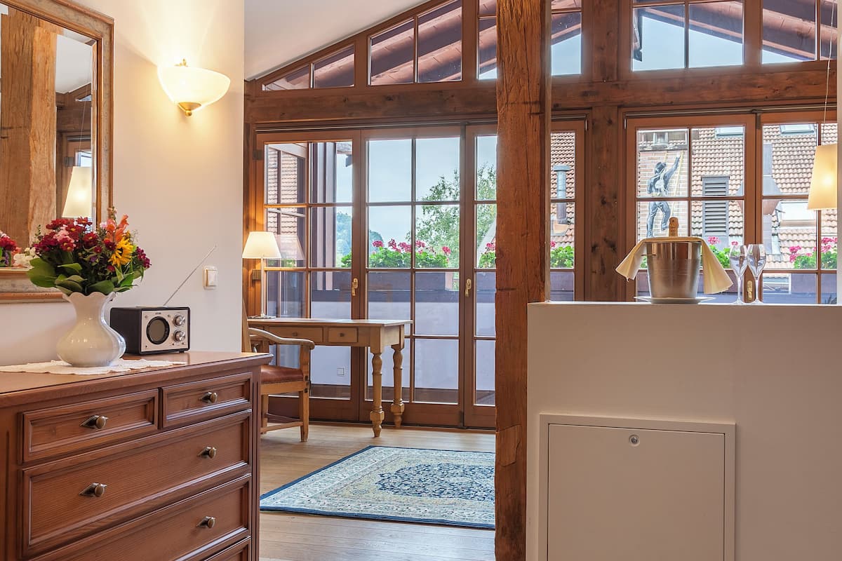 A room with wooden beams, a large window, a dresser, a radio, and a table with a lamp.