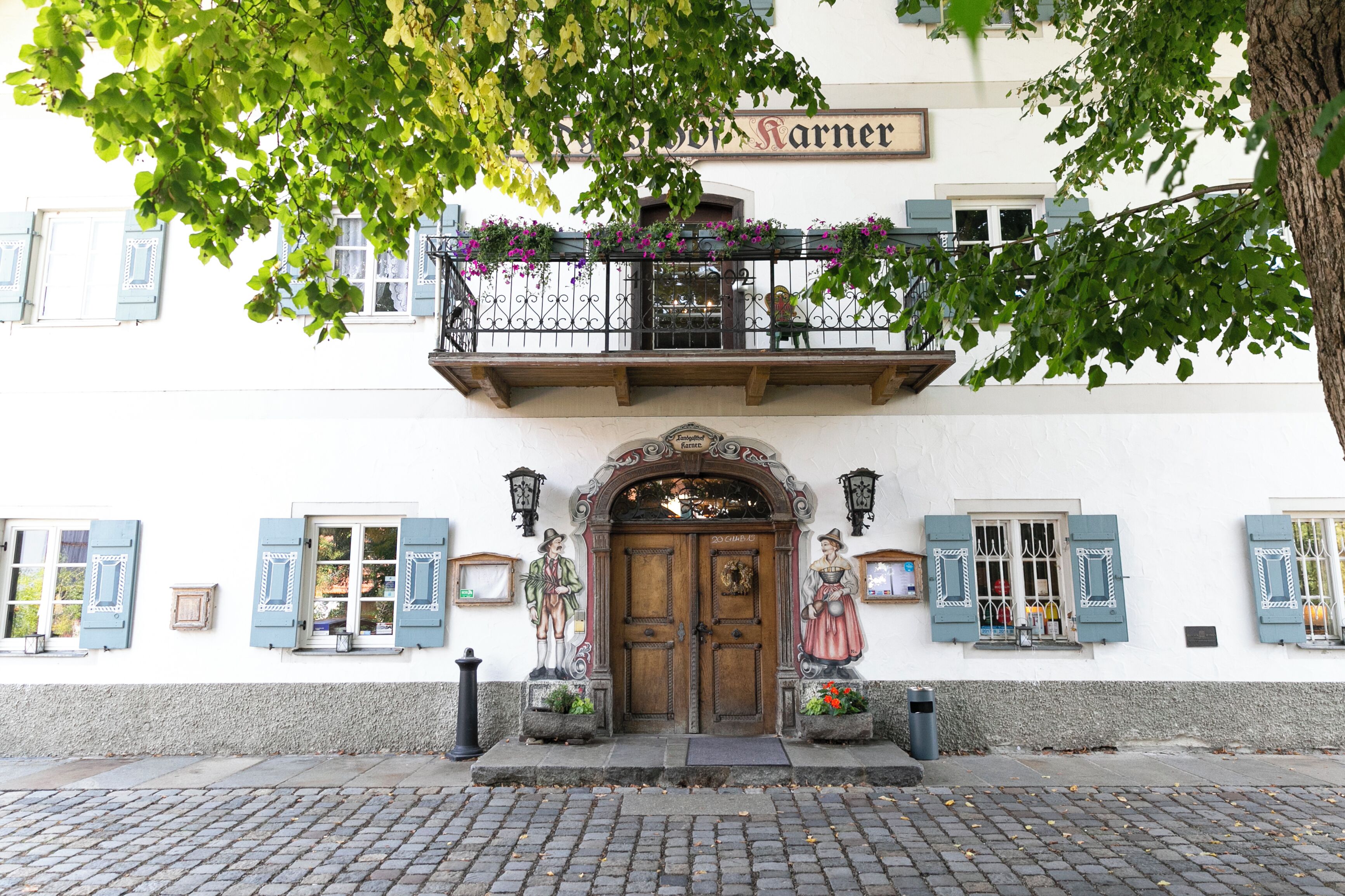 A building with a wooden double door, a balcony with flowers, and two statues on either side of the entrance.