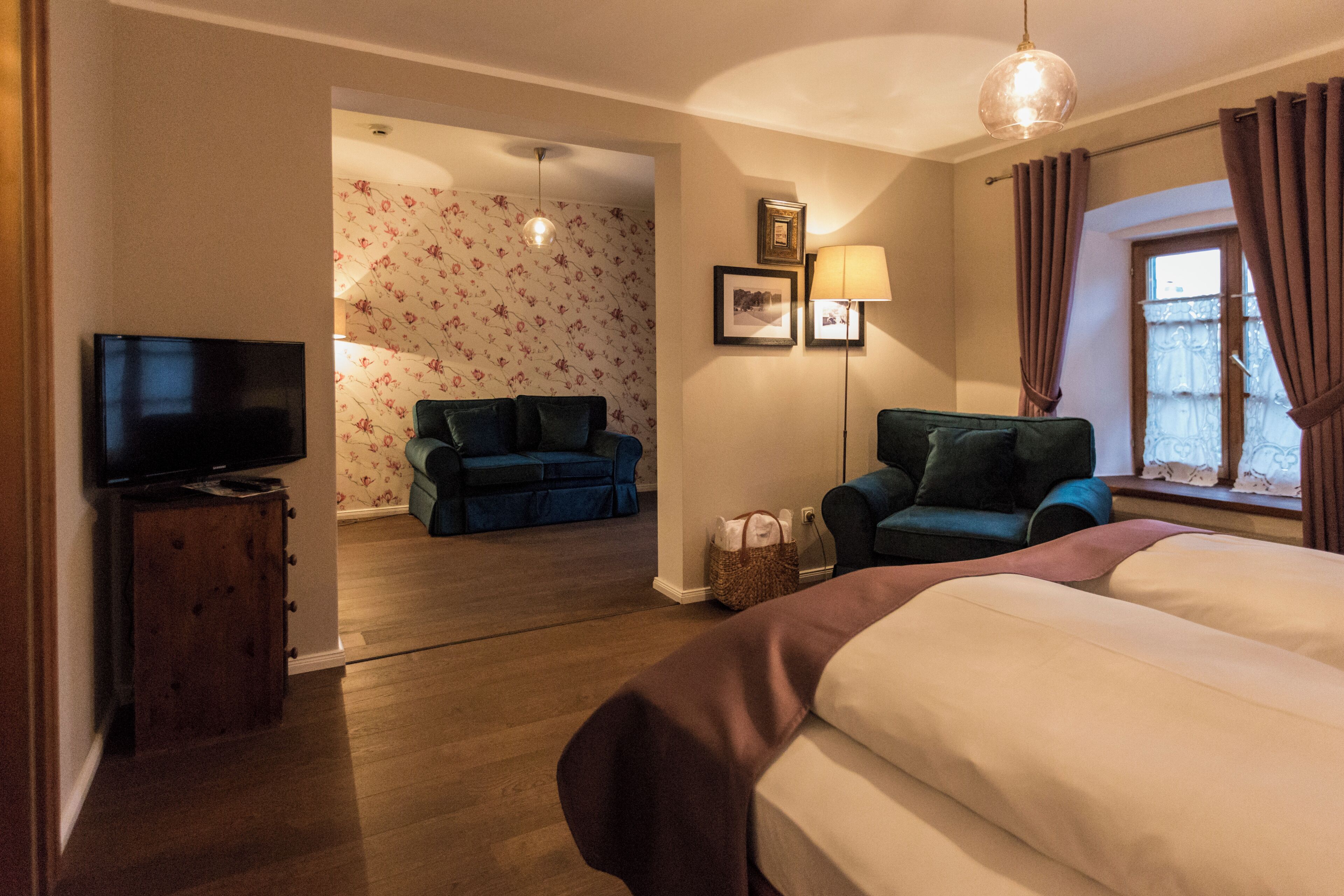 A hotel room with a bed, a television, a sofa, a chair, a lamp, and a window with curtains.