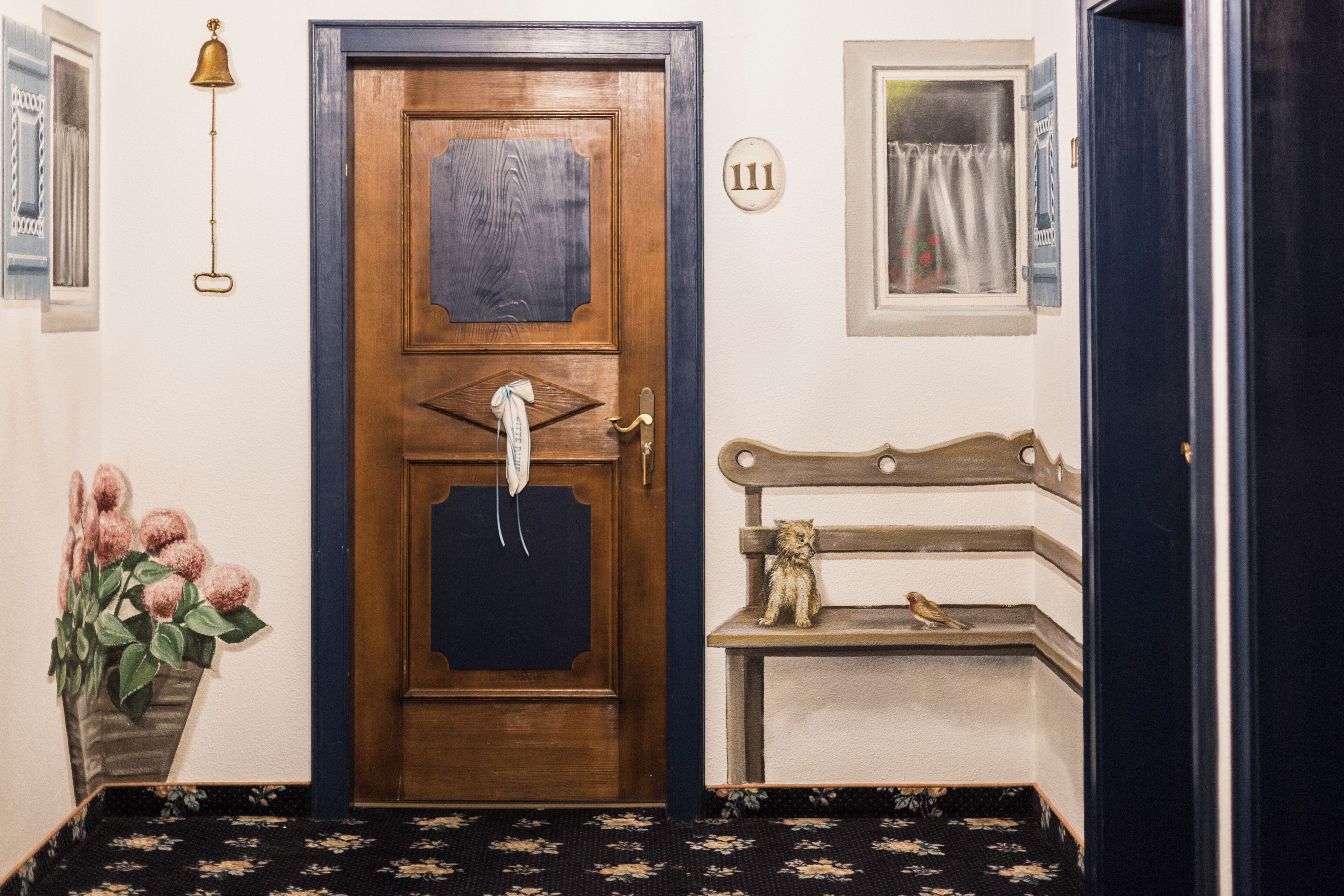 A wooden door with a number plate, a bell, a vase with flowers, and a bench with a teddy bear.