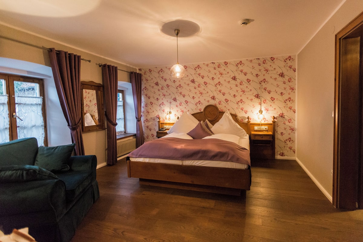 A bedroom with a bed, bedside tables, a window with curtains, a sofa, and a floral wallpaper pattern.