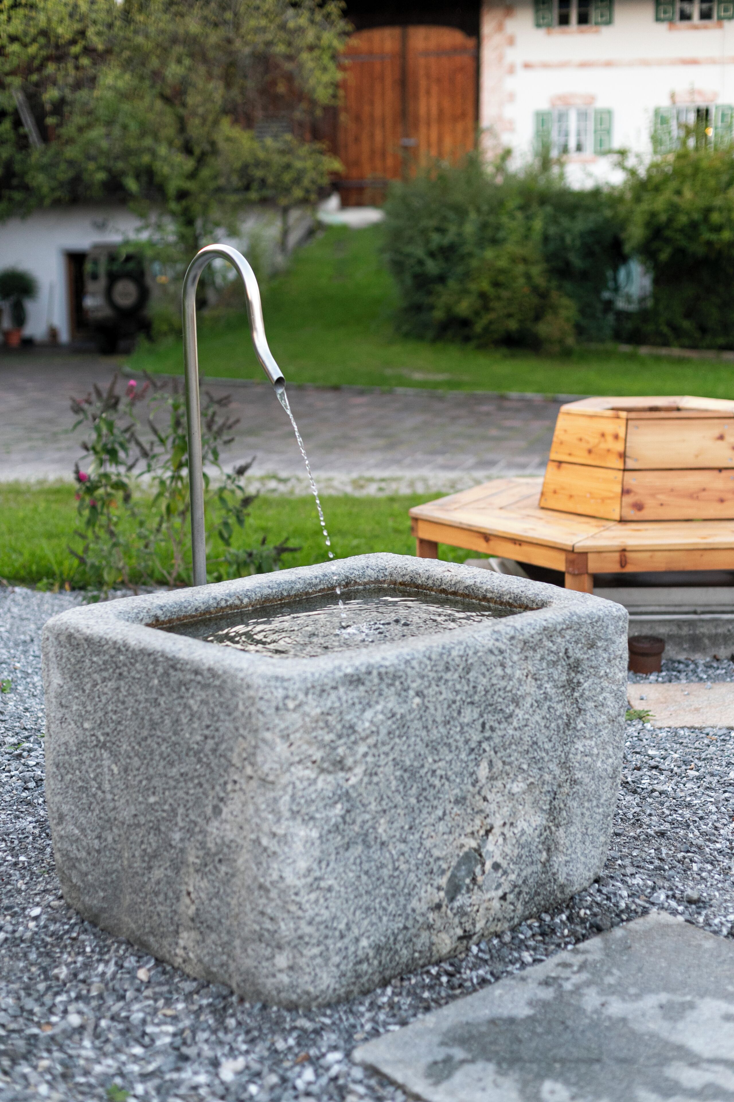Une fontaine en pierre avec un bec verseur en métal, entourée de gravier, avec un banc en bois et un bâtiment en arrière-plan.