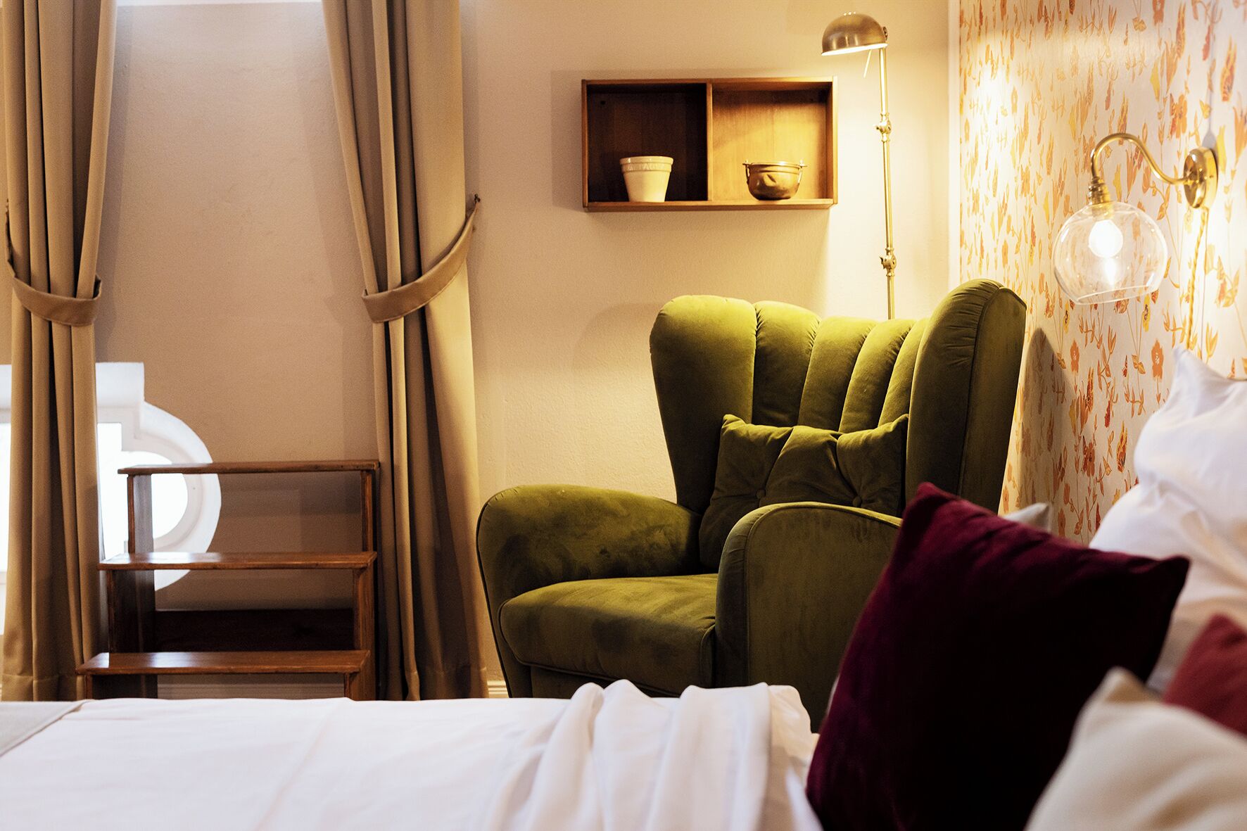 A hotel room with a green armchair, a bedside table, a lamp, and a shelf with decorative items.