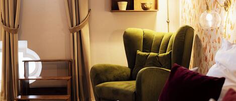A hotel room with a green armchair, a bedside table, a lamp, and a shelf with decorative items.