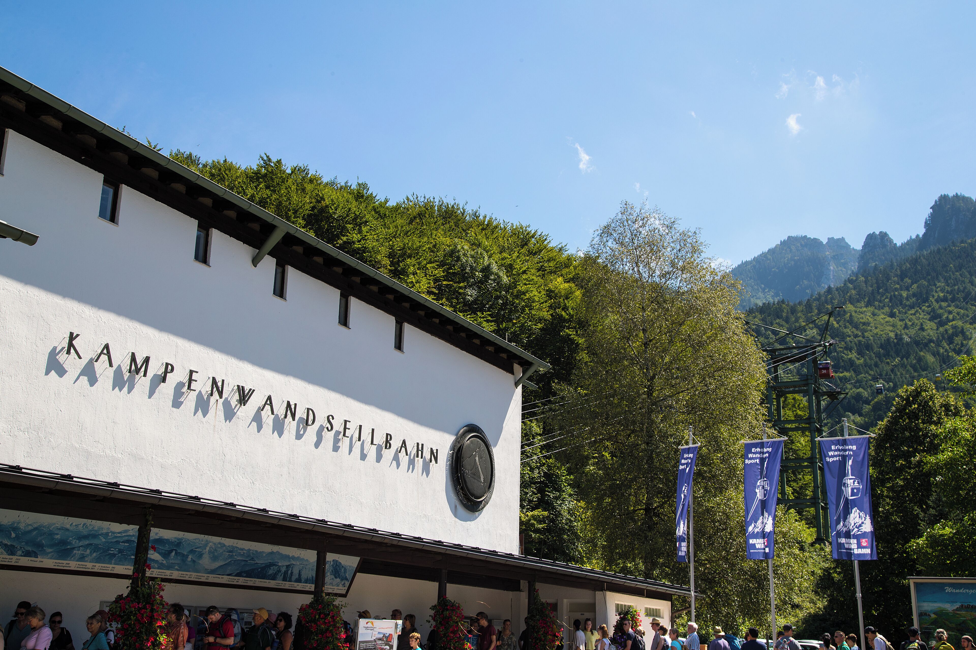 A building with 'KAMPENWANDSEILBAHN' signage, a clock, and a mountain view.