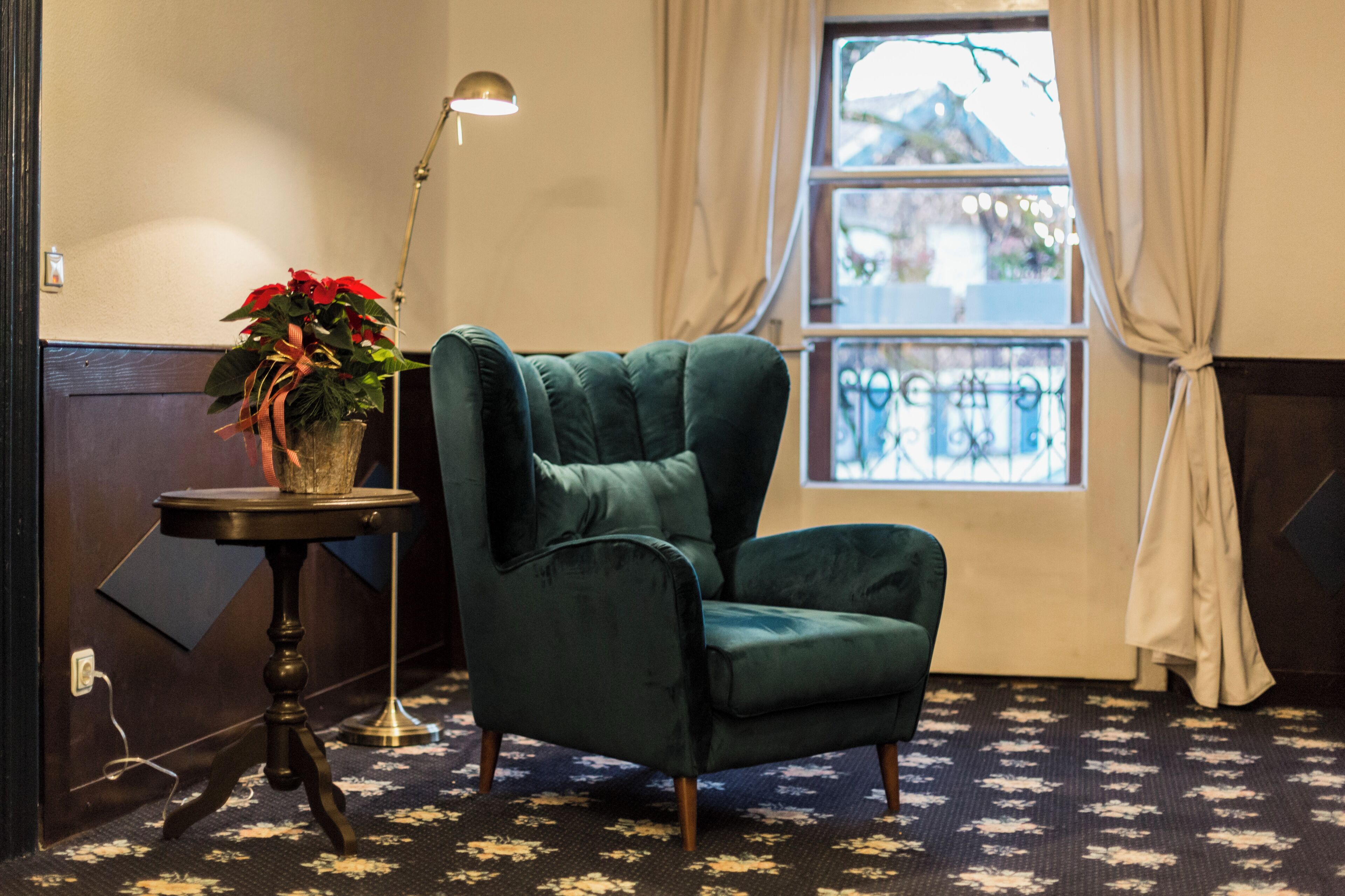 A room with a green armchair, a small table with a potted plant, a floor lamp, and a window with curtains.