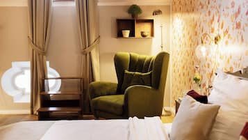 A hotel room with a bed, a green armchair, a wooden side table, a floor lamp, and a decorative wall with floral wallpaper.