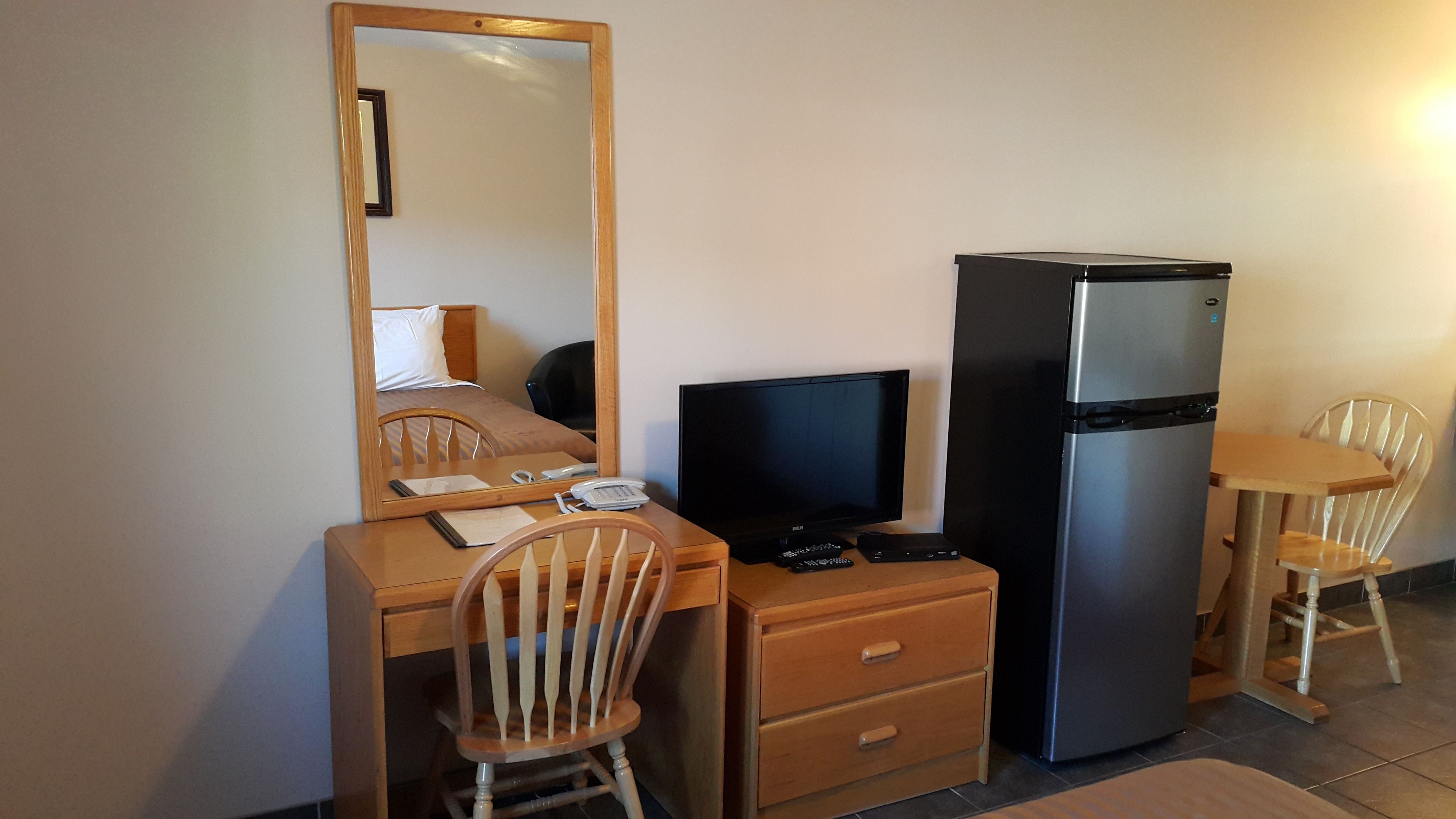 Une chambre d’hôtel de taille modeste, équipée d’un bureau en bois, d’un ordinateur, d’un réfrigérateur, d’une chaise et d’un miroir.