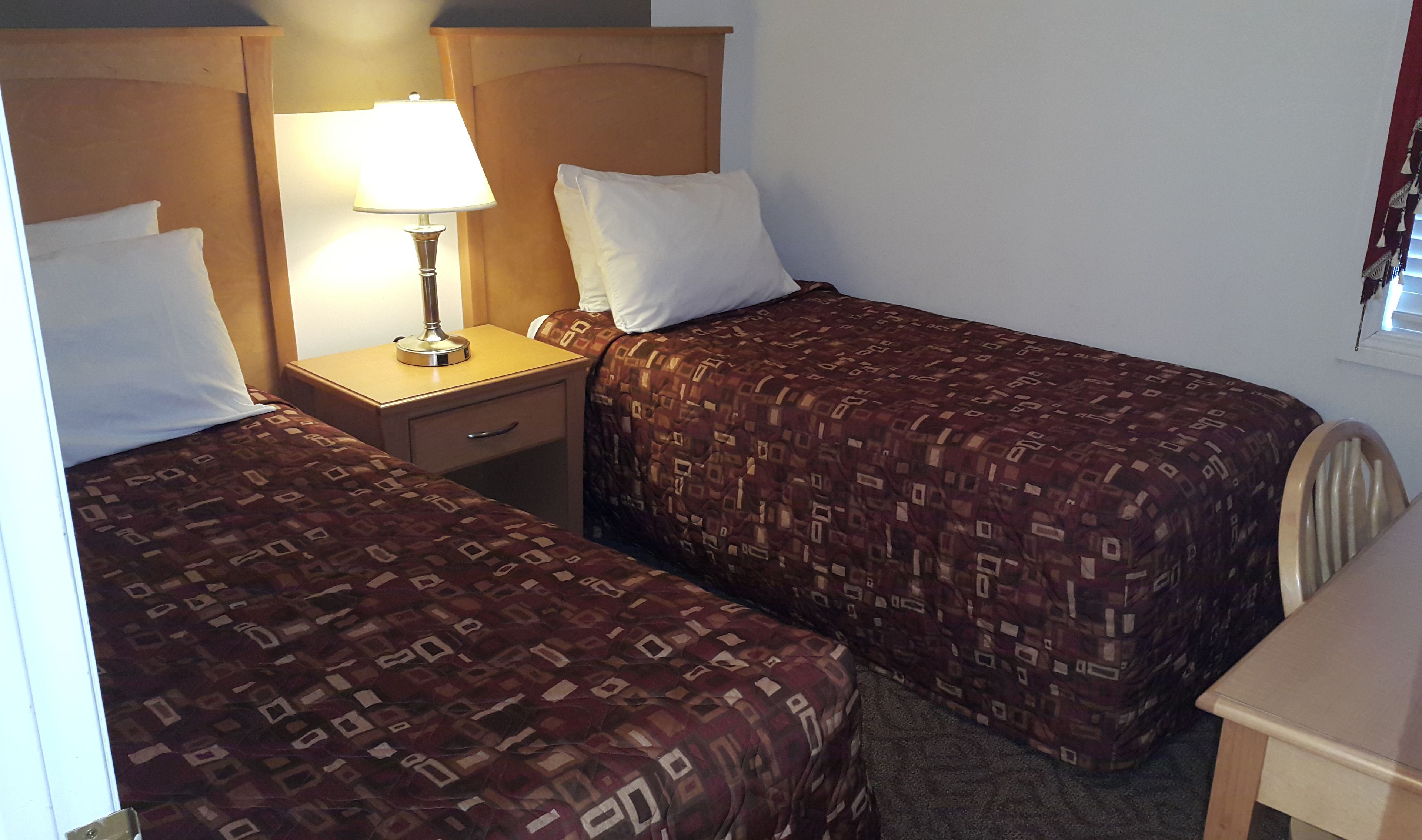 Ein Hotelzimmer mit zwei Einzelbetten, einem Nachttisch mit Lampe, einer kleinen Schublade und einem Fenster mit Vorhängen.