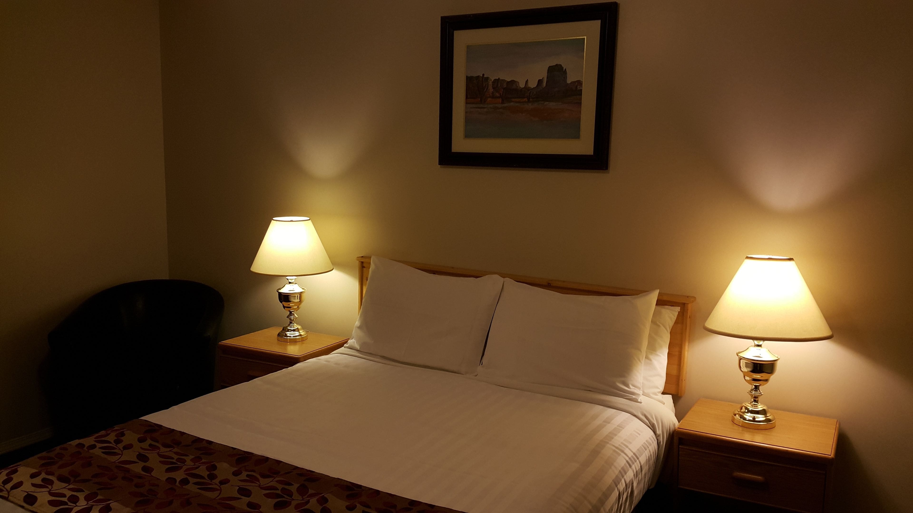A hotel room with a bed, two bedside lamps, a chair, and a framed picture on the wall.