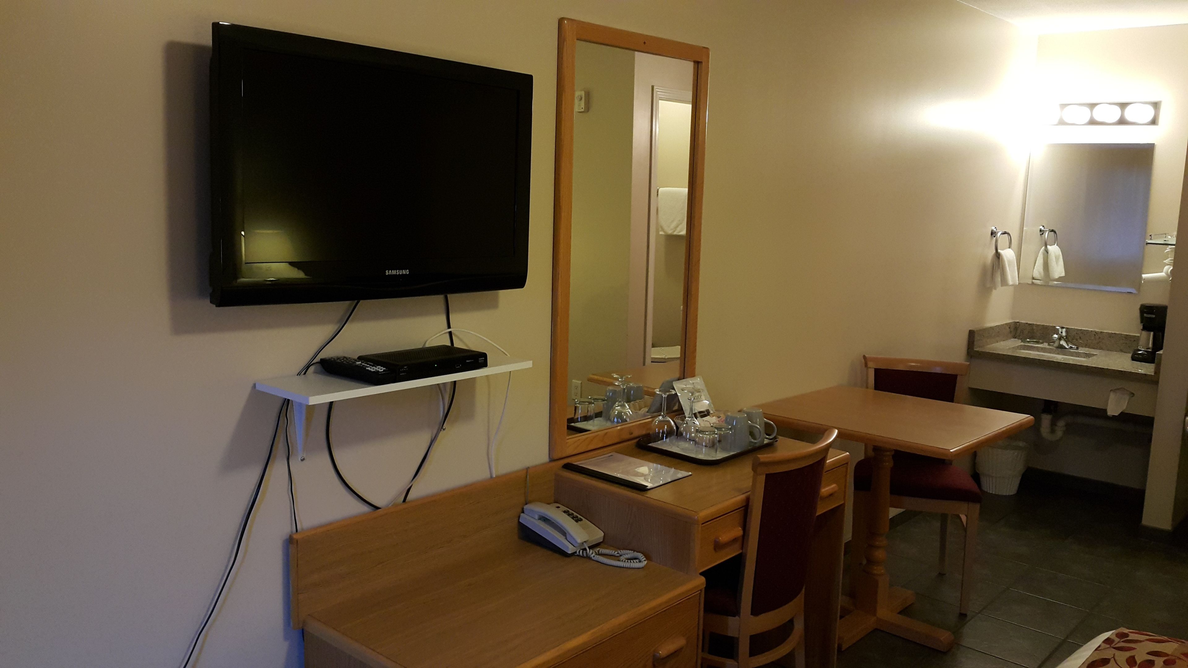 Une chambre d’hôtel équipée d’une télévision, d’un bureau avec un téléphone, d’un miroir, d’une petite table avec des verres et un plateau, ainsi que d’une salle de bain avec un lavabo et un miroir.