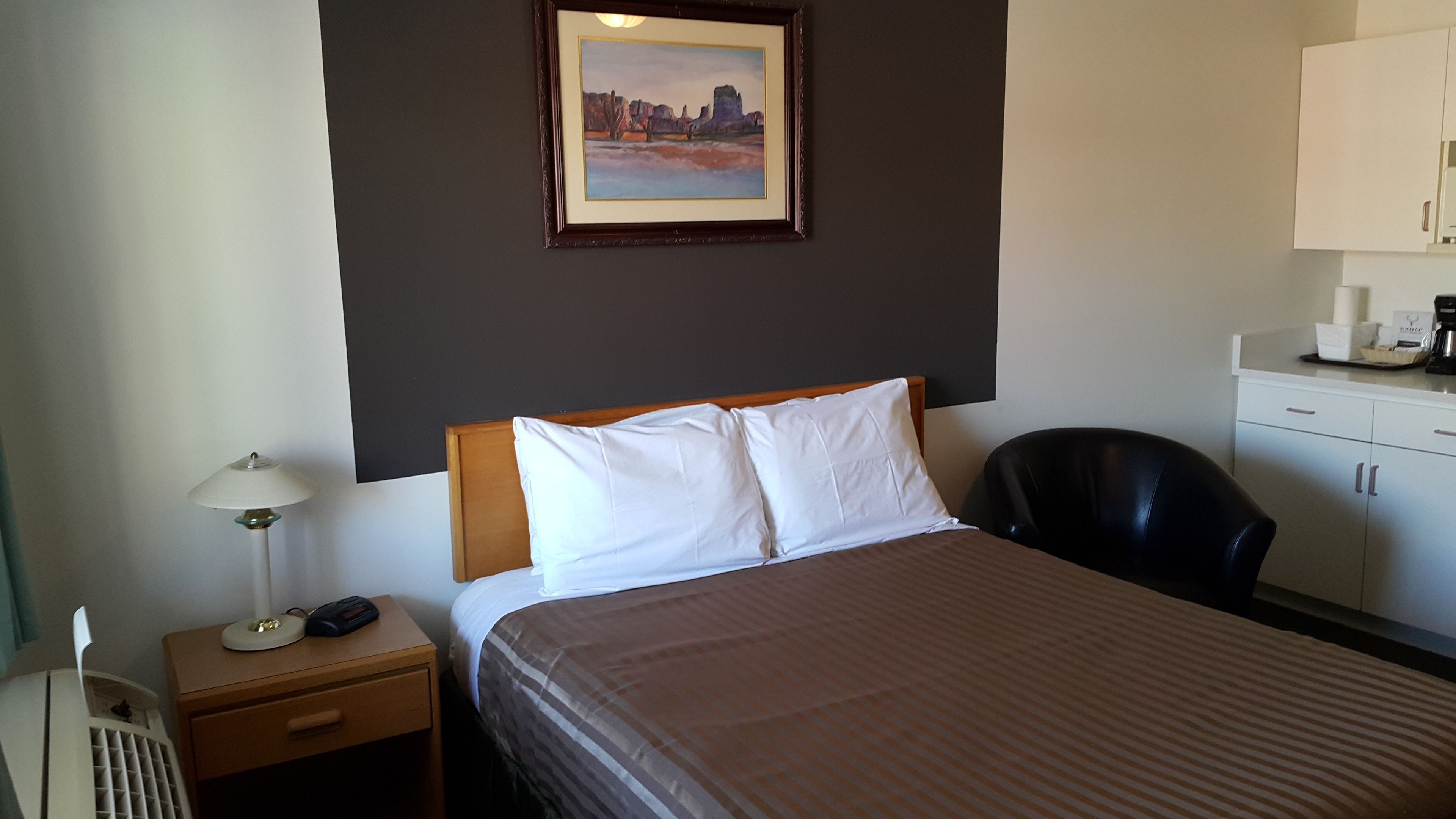 A hotel room with a bed, a nightstand with a lamp, a black chair, a painting on the wall, and a kitchenette area.
