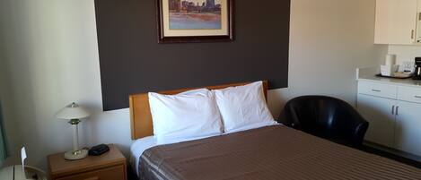 A hotel room with a bed, a nightstand with a lamp, a black chair, a painting on the wall, and a kitchenette area.