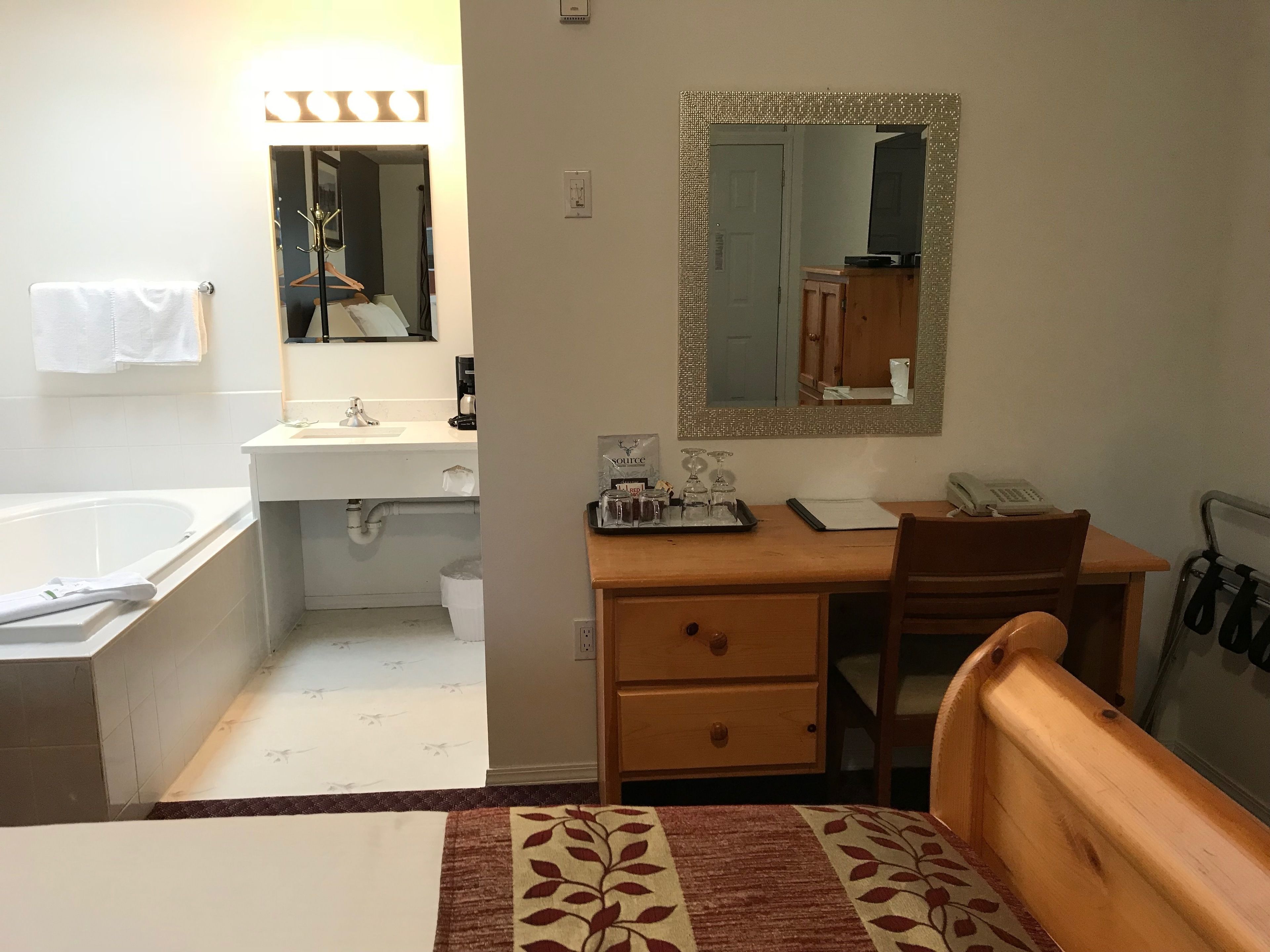 Une chambre d’hôtel comprenant une salle de bain, un bureau en bois avec une chaise, un lit recouvert d’une courtepointe à motifs et un miroir avec un cadre décoratif.