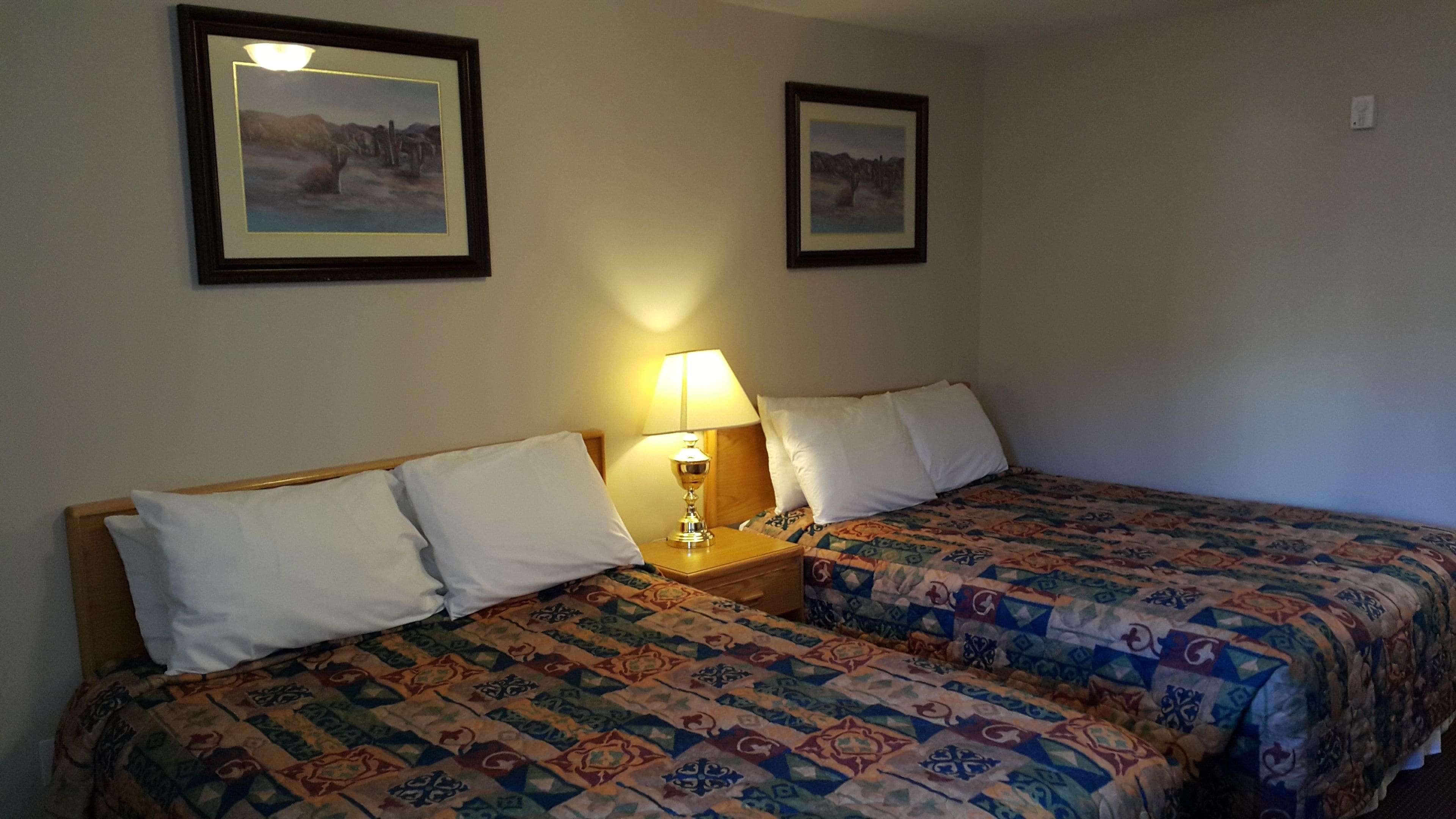 Une chambre avec deux lits, chacun recouvert d’une couvre-lit à motifs, une table de chevet avec une lampe, et deux tableaux encadrés accrochés au mur.