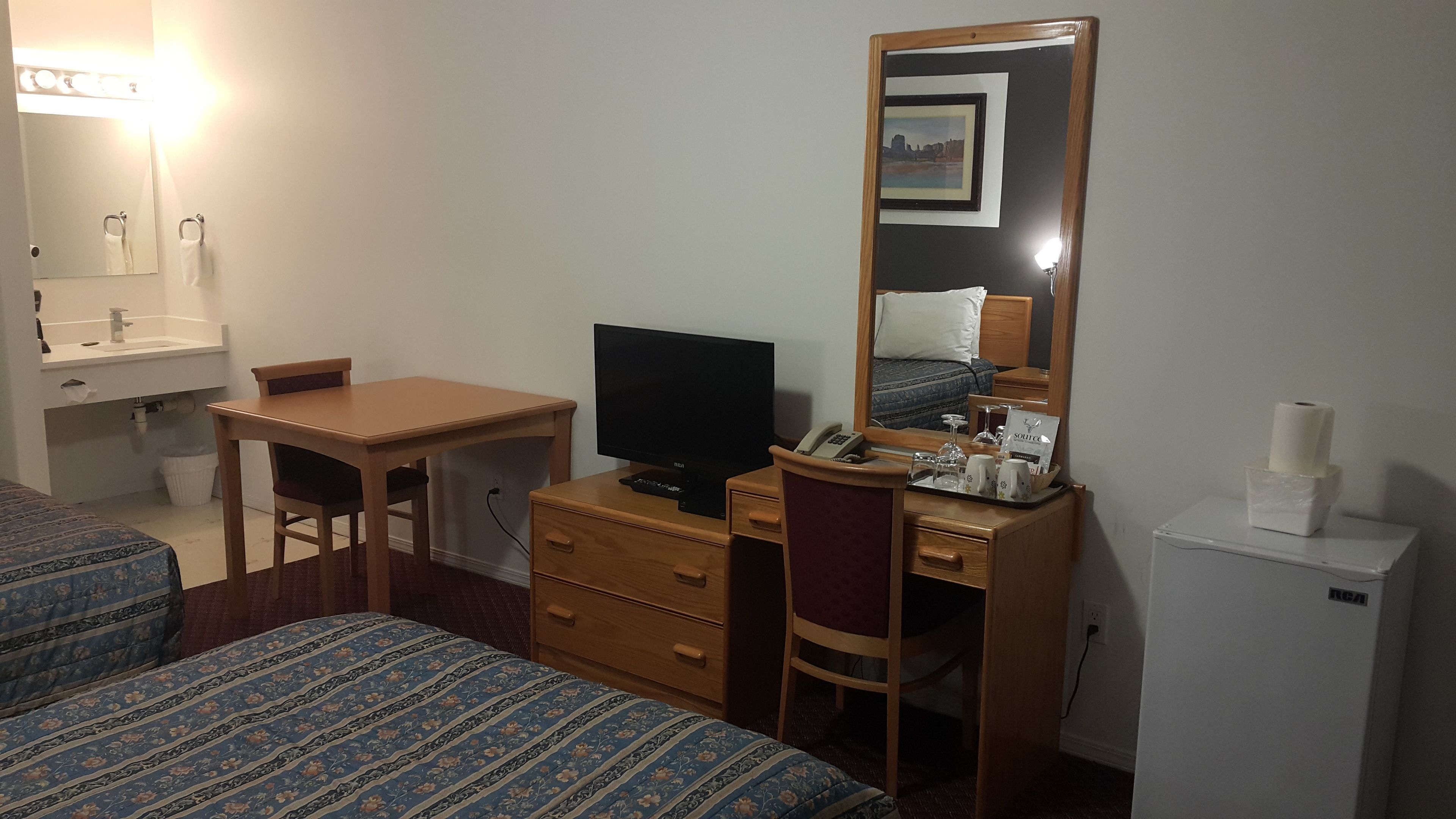 Habitación de hotel con cama, escritorio con televisor, espejo, refrigerador pequeño y mesita de noche con lámpara y una bandeja con objetos.