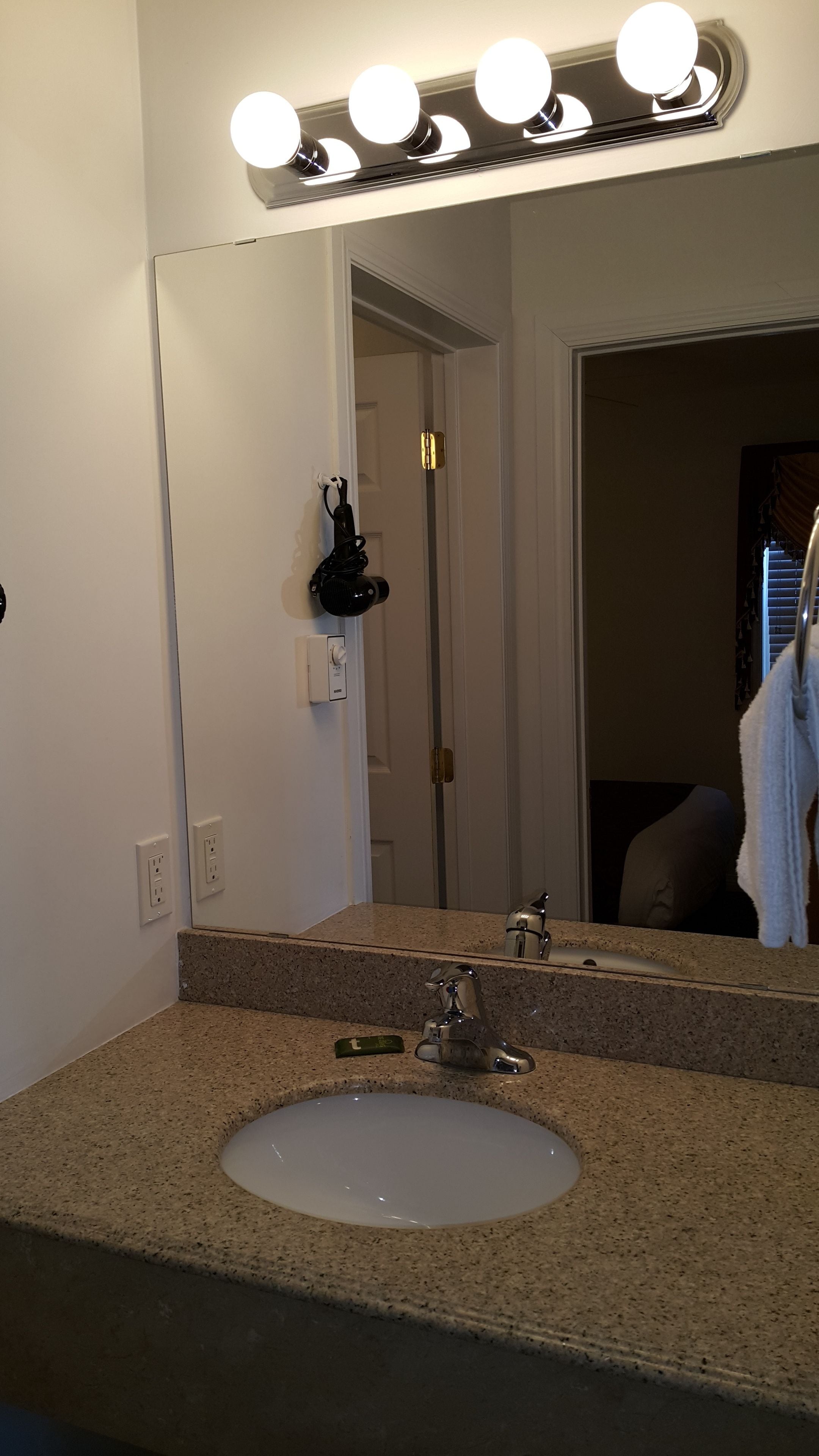 Un baño con encimera de granito, un lavamanos blanco, un espejo con iluminación, un perchero y una puerta que da a otra habitación.