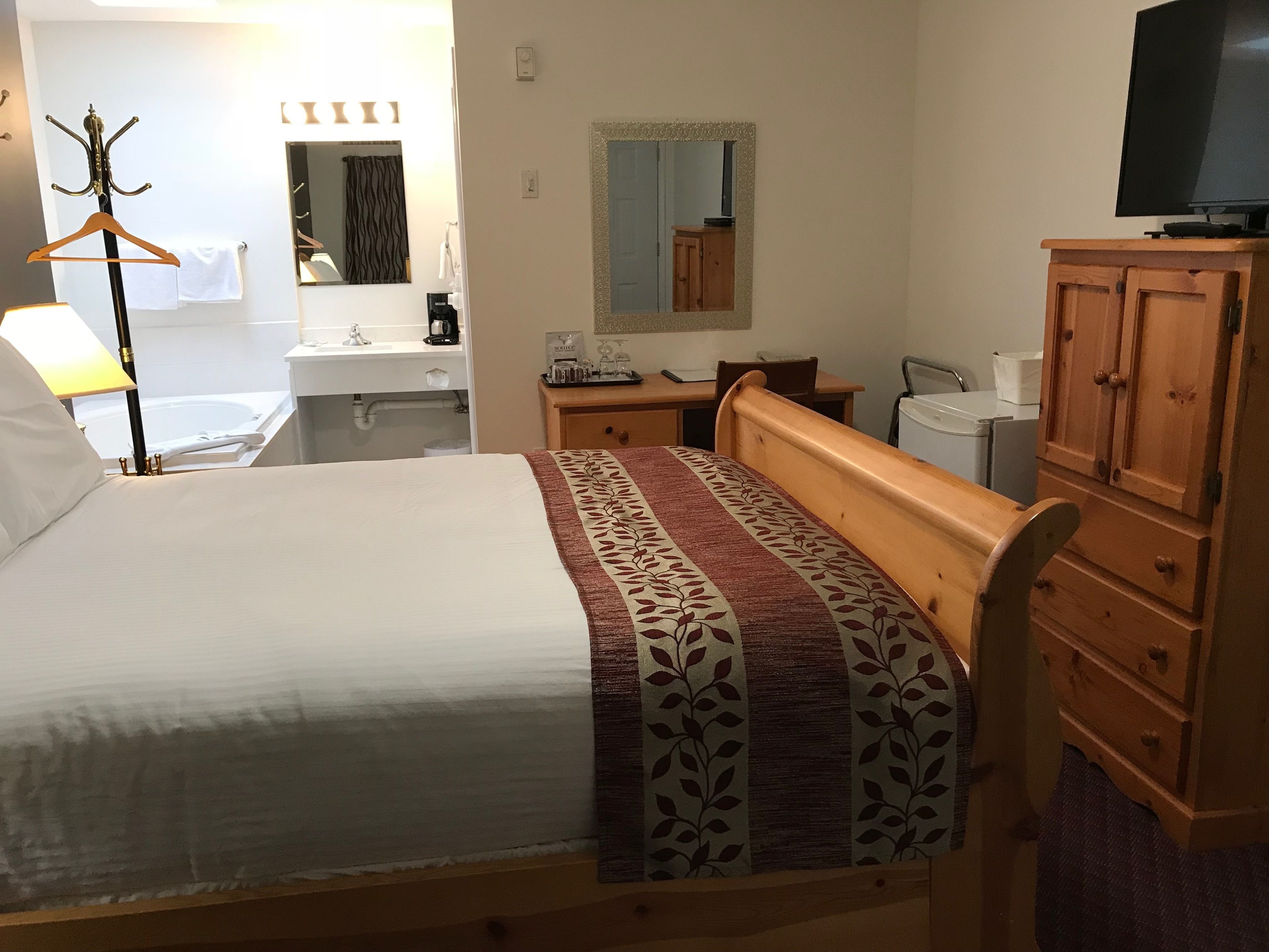 A hotel room with a bed, a wooden dresser, a television, a desk with a chair, a mirror, and a bathroom with a sink and toilet.