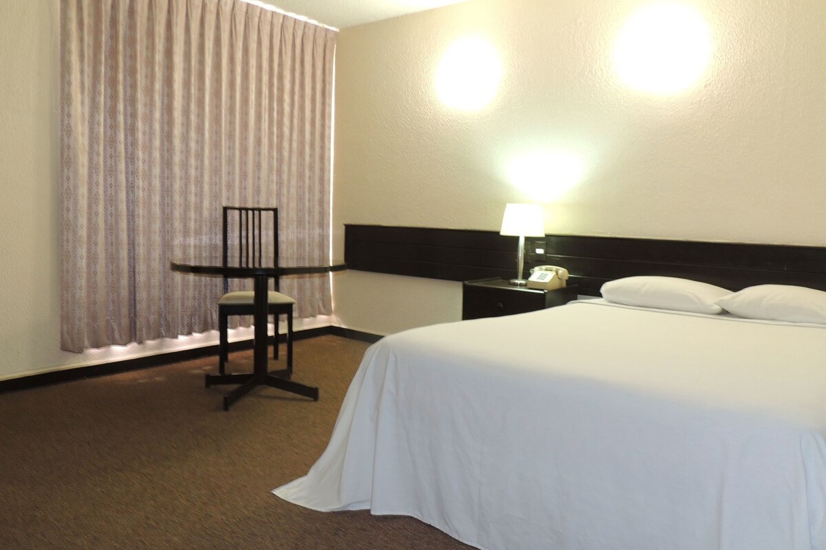 A hotel room with a bed, a desk, a chair, a nightstand with a lamp, and a telephone.