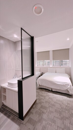 A modern hotel room with a large bed, a separate bathroom with a vanity, and a window with a view of buildings.