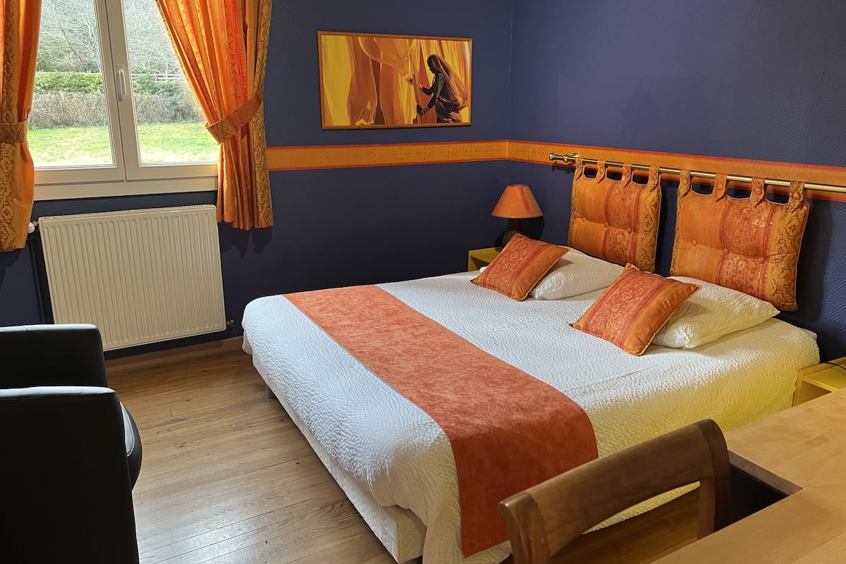 Una habitación de hotel ordenada con una cama grande, una silla, una mesa y un ventanal con cortinas.