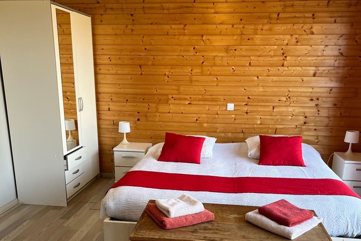Un dormitorio con techo de madera, cama con ropa de cama roja y blanca, dos mesitas de noche con lámparas y un aparador de madera.