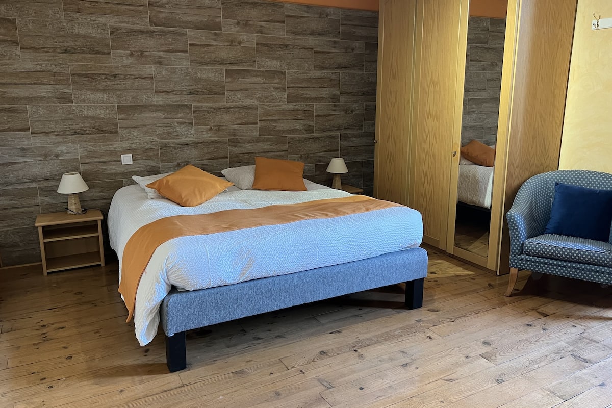 Un dormitorio con cama, dos mesitas de noche, un armario de madera y un sillón estampado.