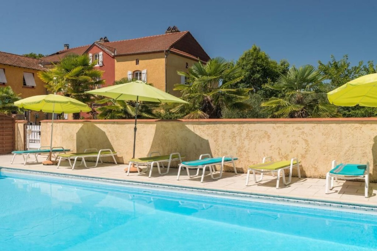 A swimming pool with lounge chairs and umbrellas, surrounded by a building with a red roof and palm trees.