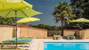 Un área junto a la piscina con sillones de descanso y sombrillas, una piscina y palmeras.