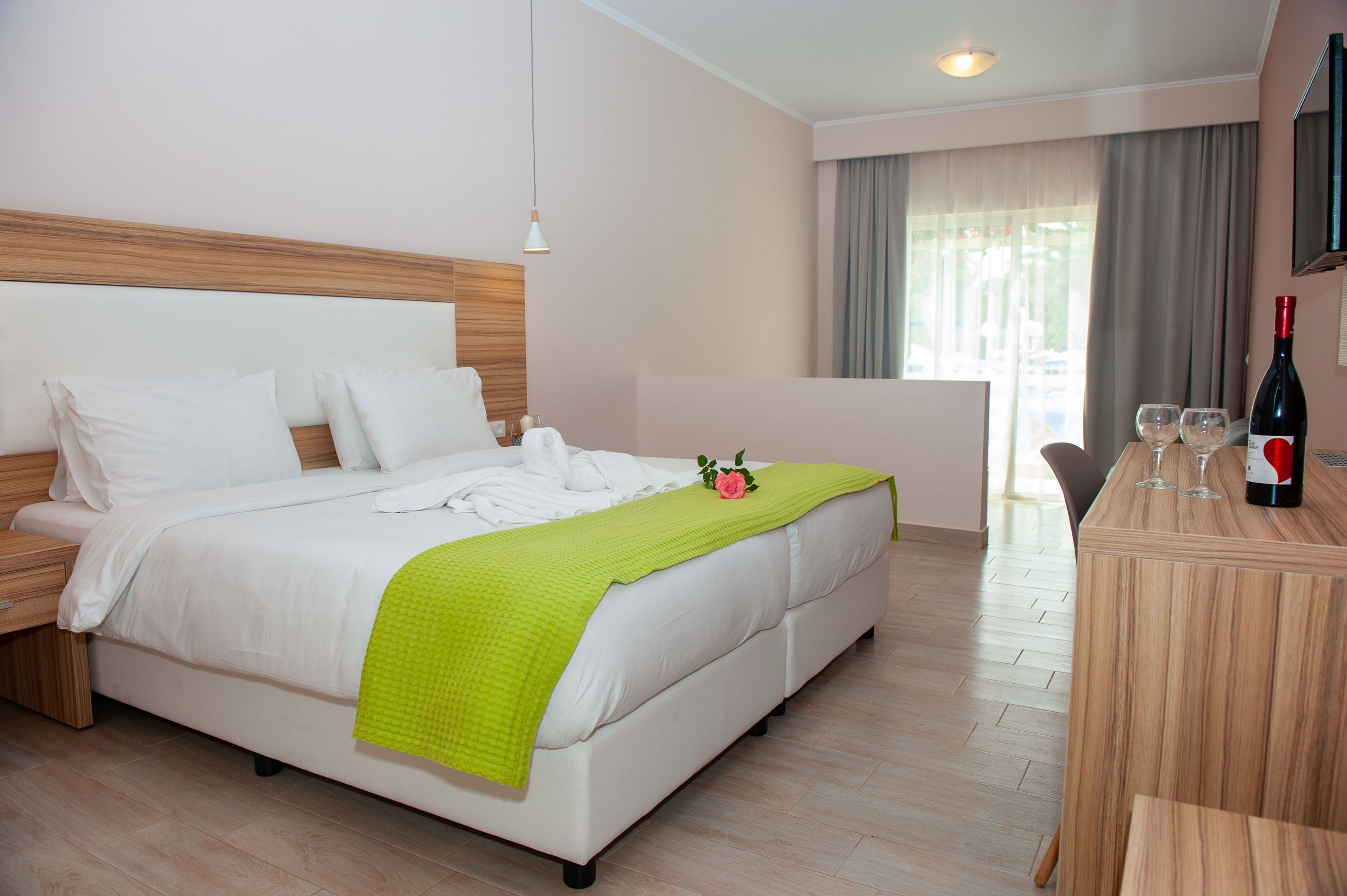 Habitación de hotel con una cama, cabecera de madera, manta verde, escritorio de madera con una botella de vino y copas, y una ventana con cortinas.