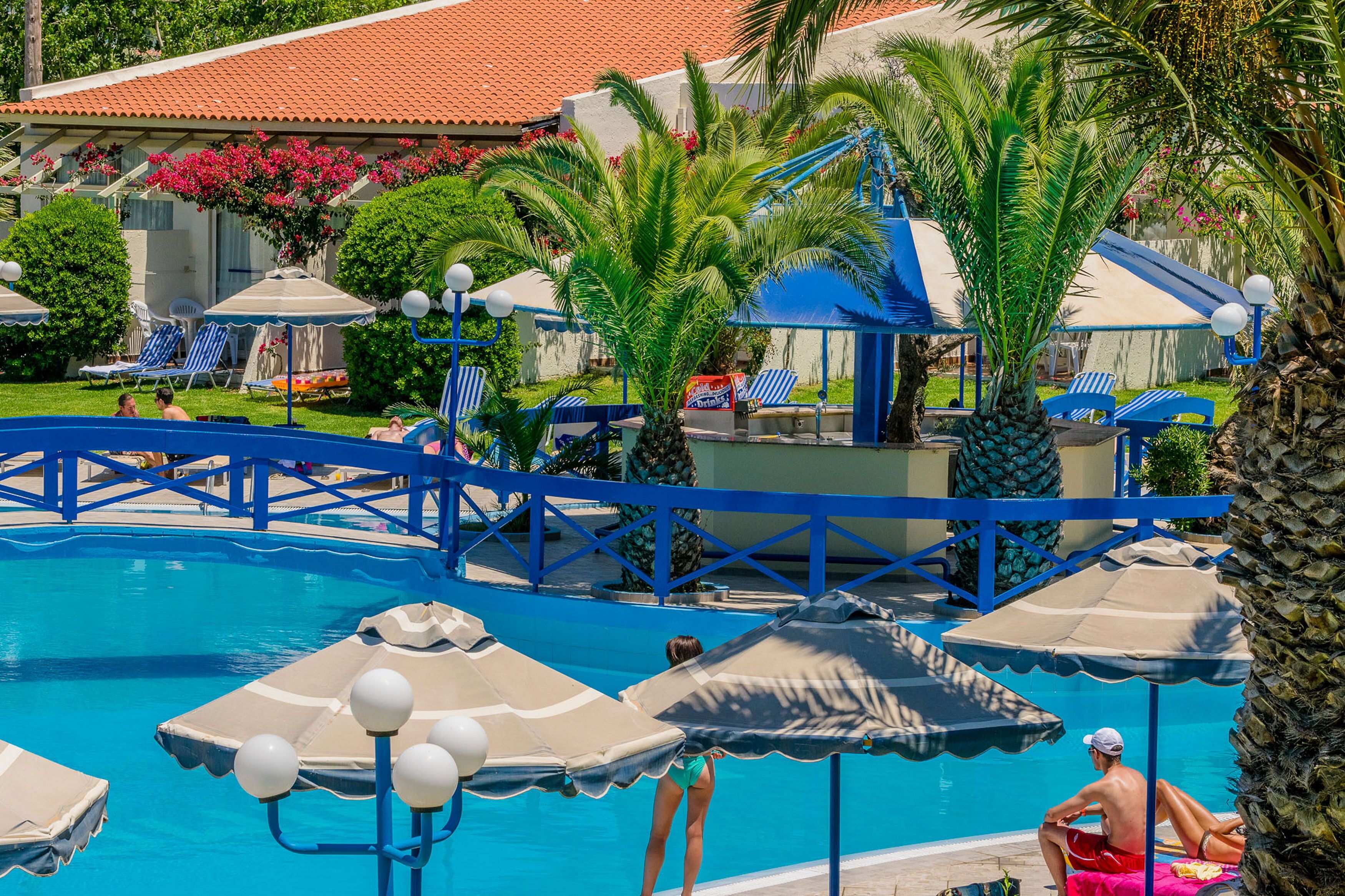 Zona de piscina con sombrillas azules y color beige, sillas de descanso y un puente sobre el agua.