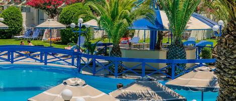 Zona de piscina con sombrillas azules y color beige, sillas de descanso y un puente sobre el agua.