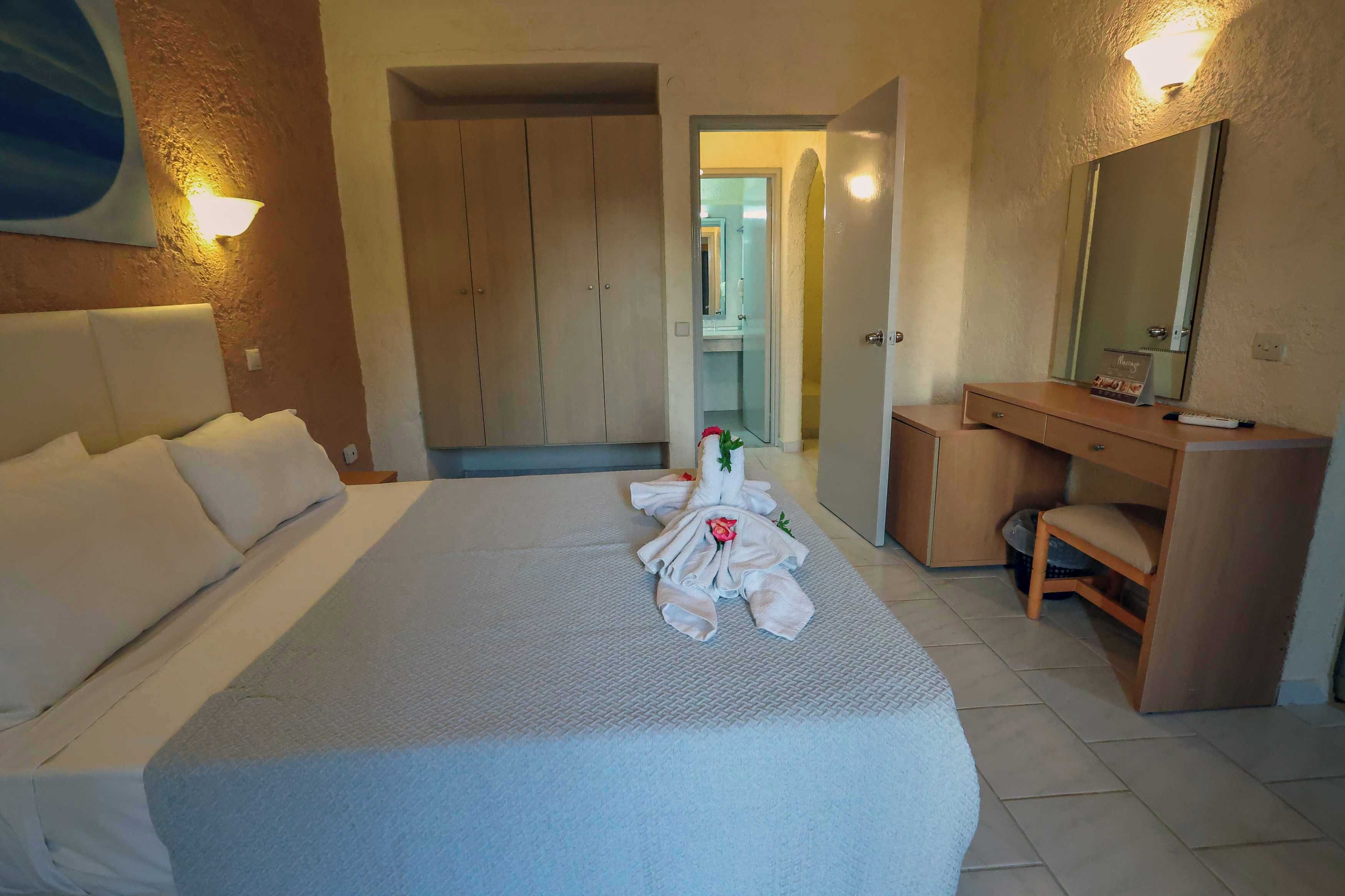 Una habitación de hotel con una cama bien hecha, un armario de madera, un escritorio pequeño con una silla y un baño visible a través de una puerta abierta.