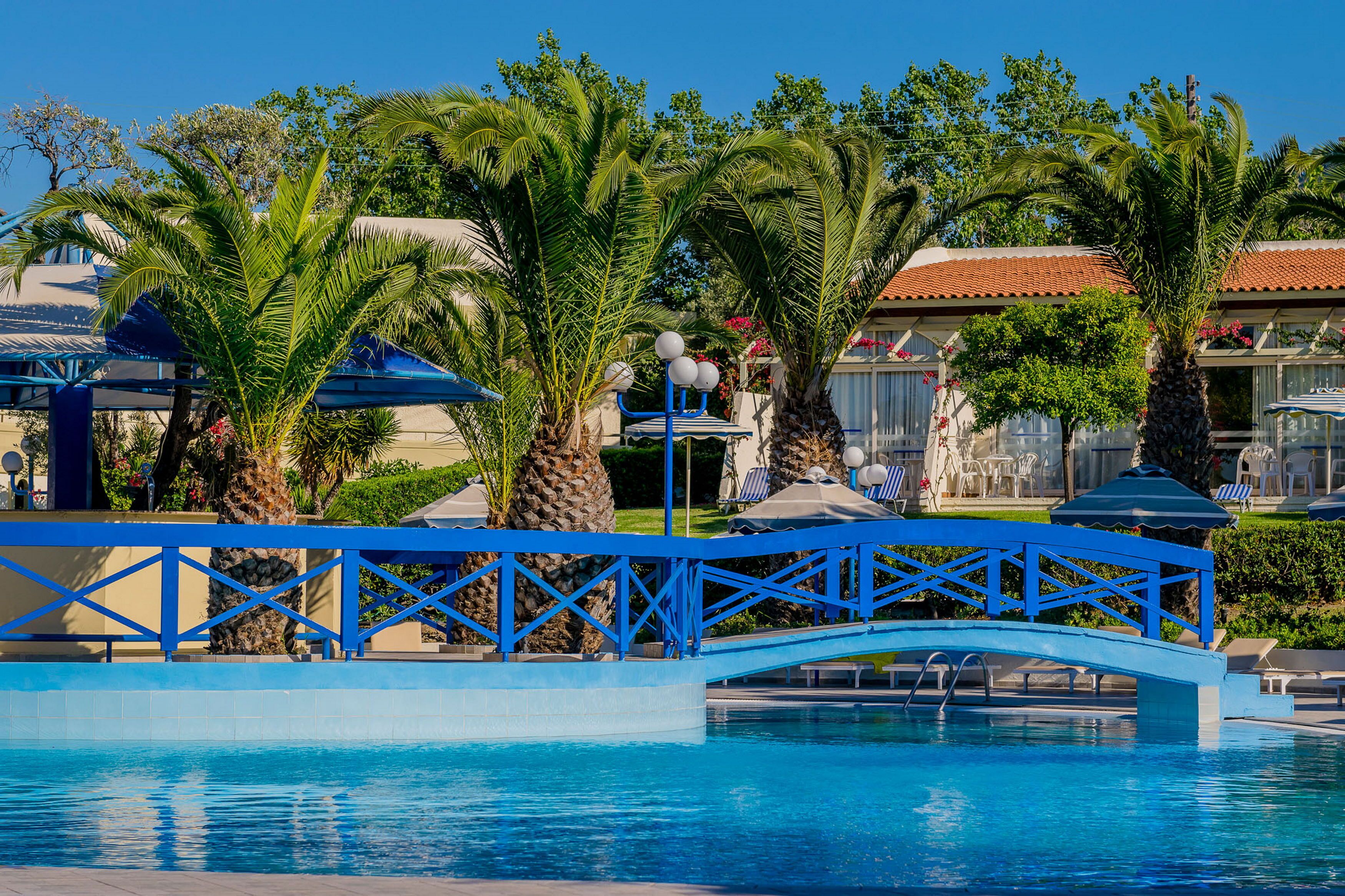 Un espace piscine agrémenté d’un pont bleu, de palmiers et de chaises longues.