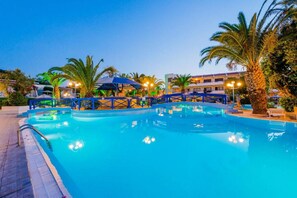 A swimming pool with a blue water feature, surrounded by palm trees and a building with balconies. - Filerimos Village - All Inclusive (Rhodes)