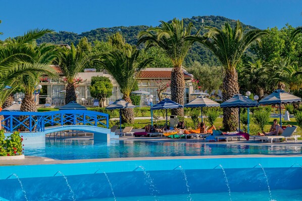 Un área de piscina con sillones de descanso y sombrillas, un puente azul y montañas al fondo.