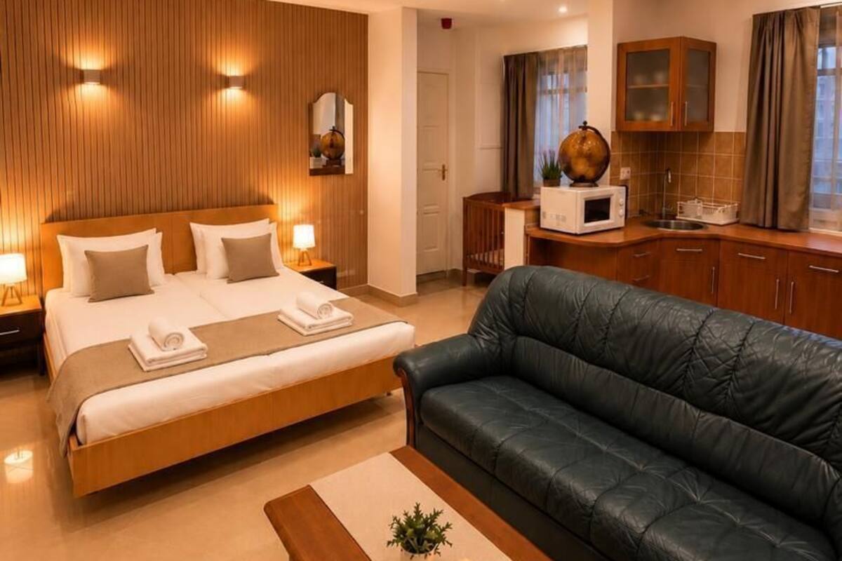 A modern hotel room with a large bed, a leather sofa, a wooden coffee table, and a kitchen area.