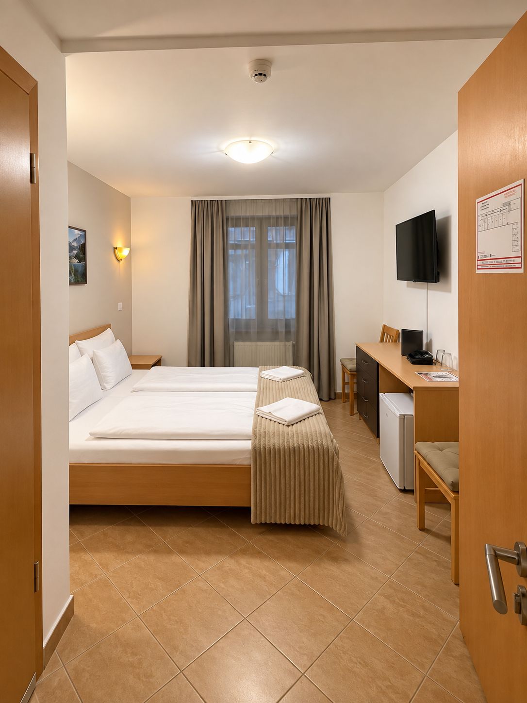 A hotel room with a bed, bedside table, desk, chair, television, and a window with curtains.