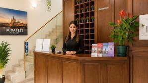A reception desk with a woman standing behind it, a staircase with a view of Budapest, and a reception area with wooden paneling and a potted plant. - Esprit Hotel Budapest (Budapest)