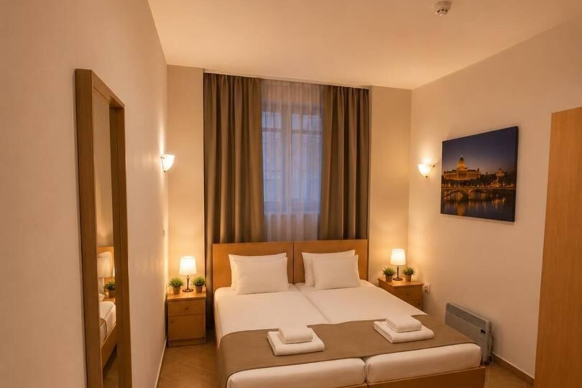 A hotel room with a large bed, bedside tables, a desk, a chair, a mirror, and a window with curtains.
