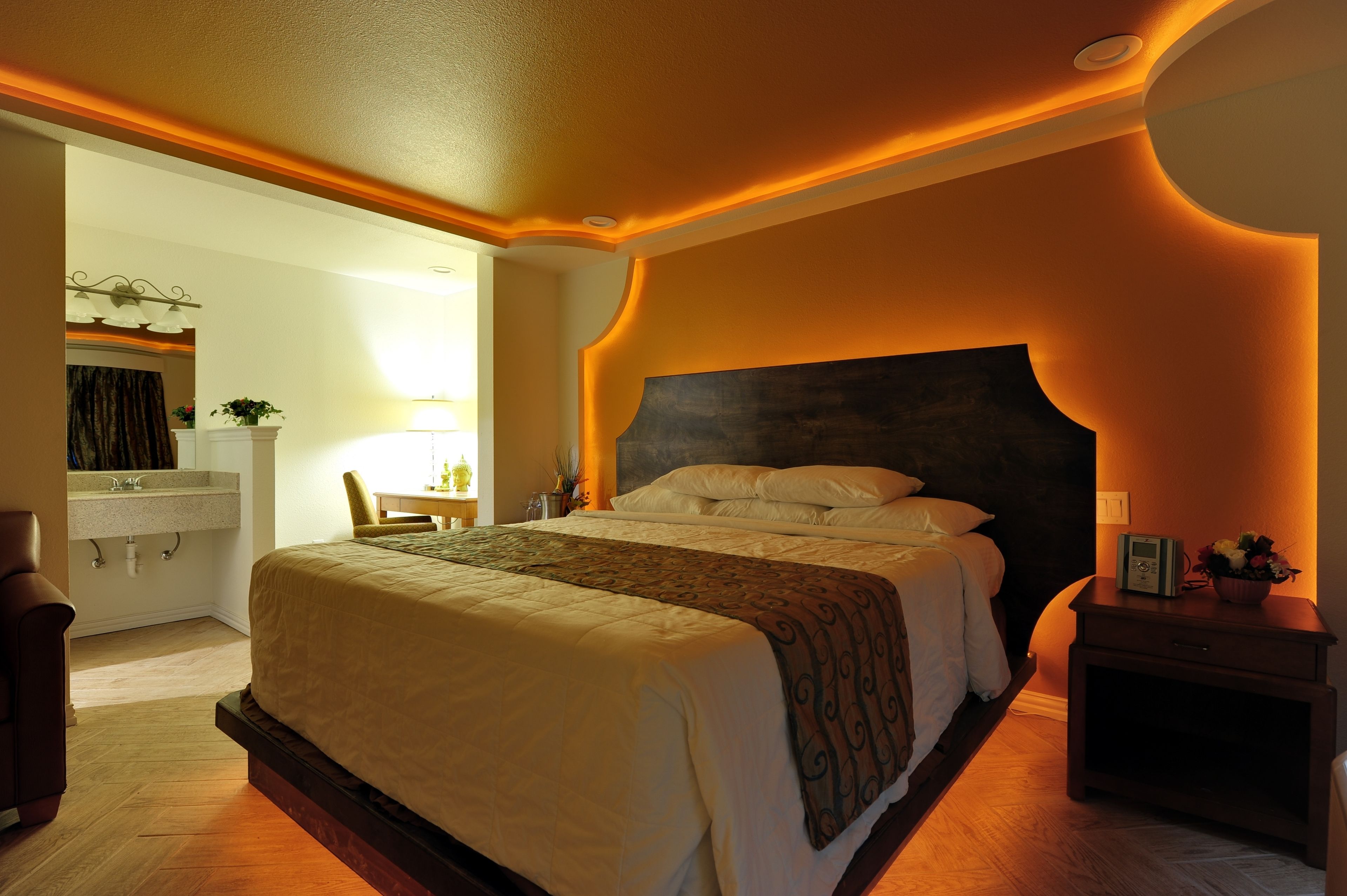 A hotel room with a large bed, a bedside table, a nightstand, a mirror, and a bathroom with a sink and towel rack.
