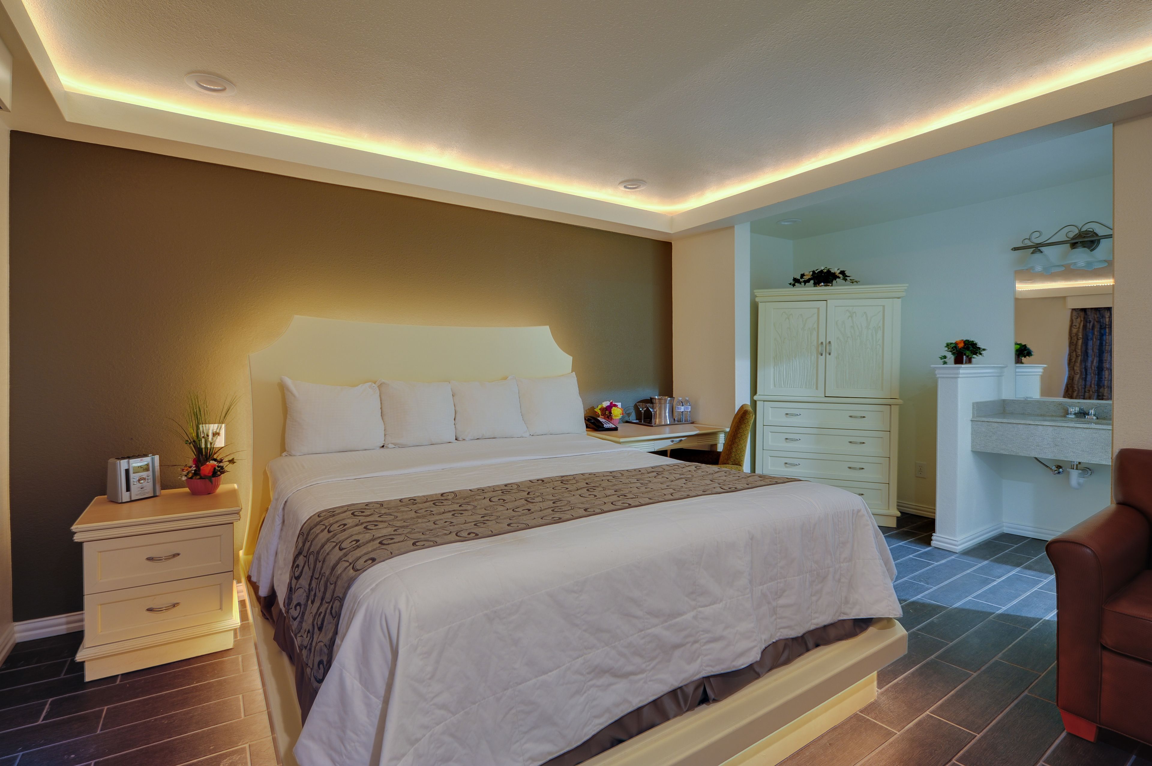 A hotel room with a large bed, bedside table, and a desk with a chair. There is a wardrobe and a bathroom visible in the background.