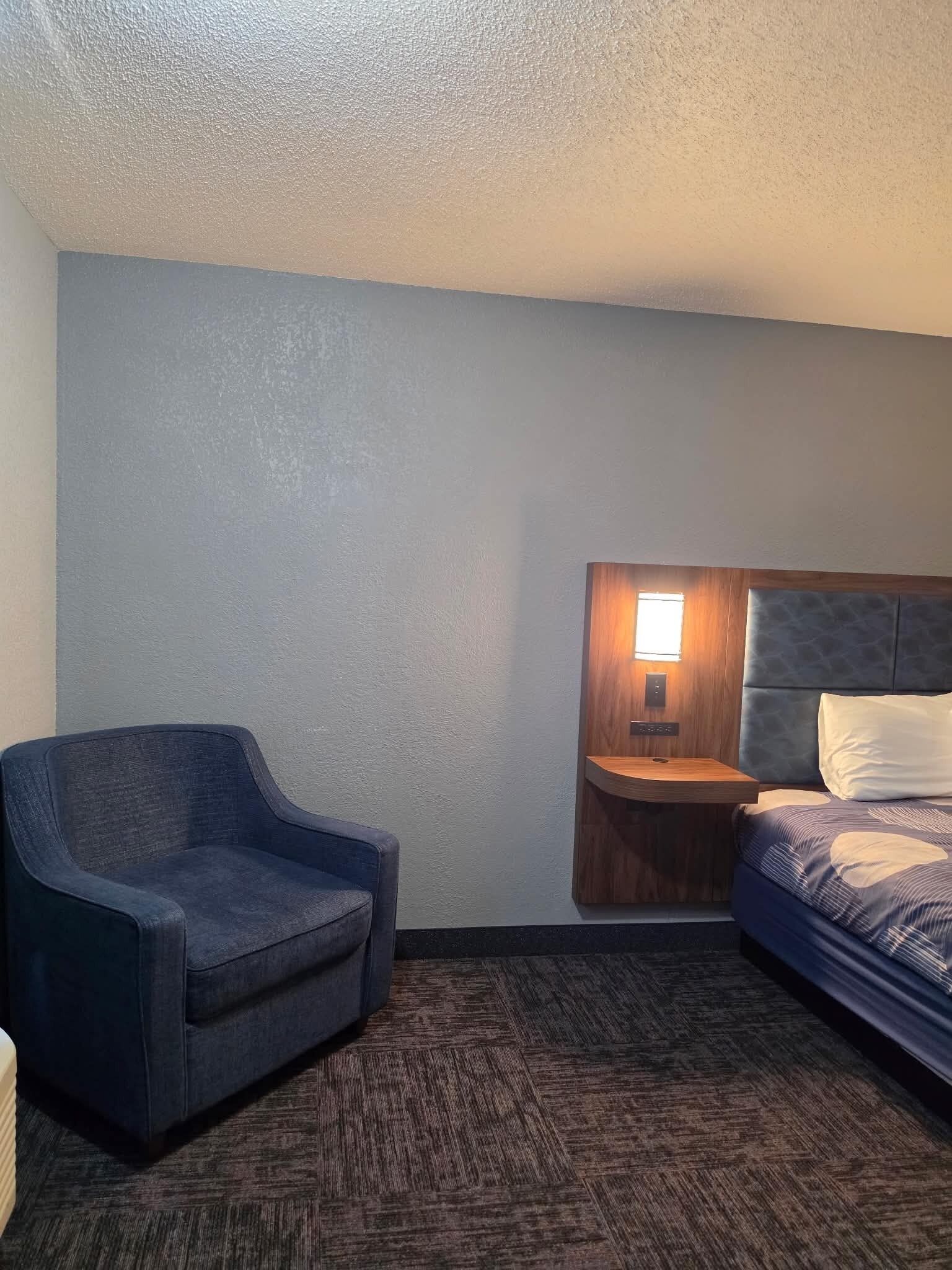 A hotel room with a dark blue armchair, a bed with a patterned bedspread, and a wooden nightstand with a built-in light.
