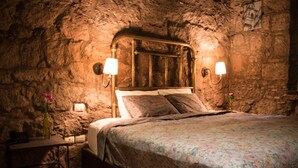 Un dormitorio con pared de piedra, cabecera de madera, dos lámparas de noche, colcha estampada con motivos florales y una mesita de noche con un jarrón.