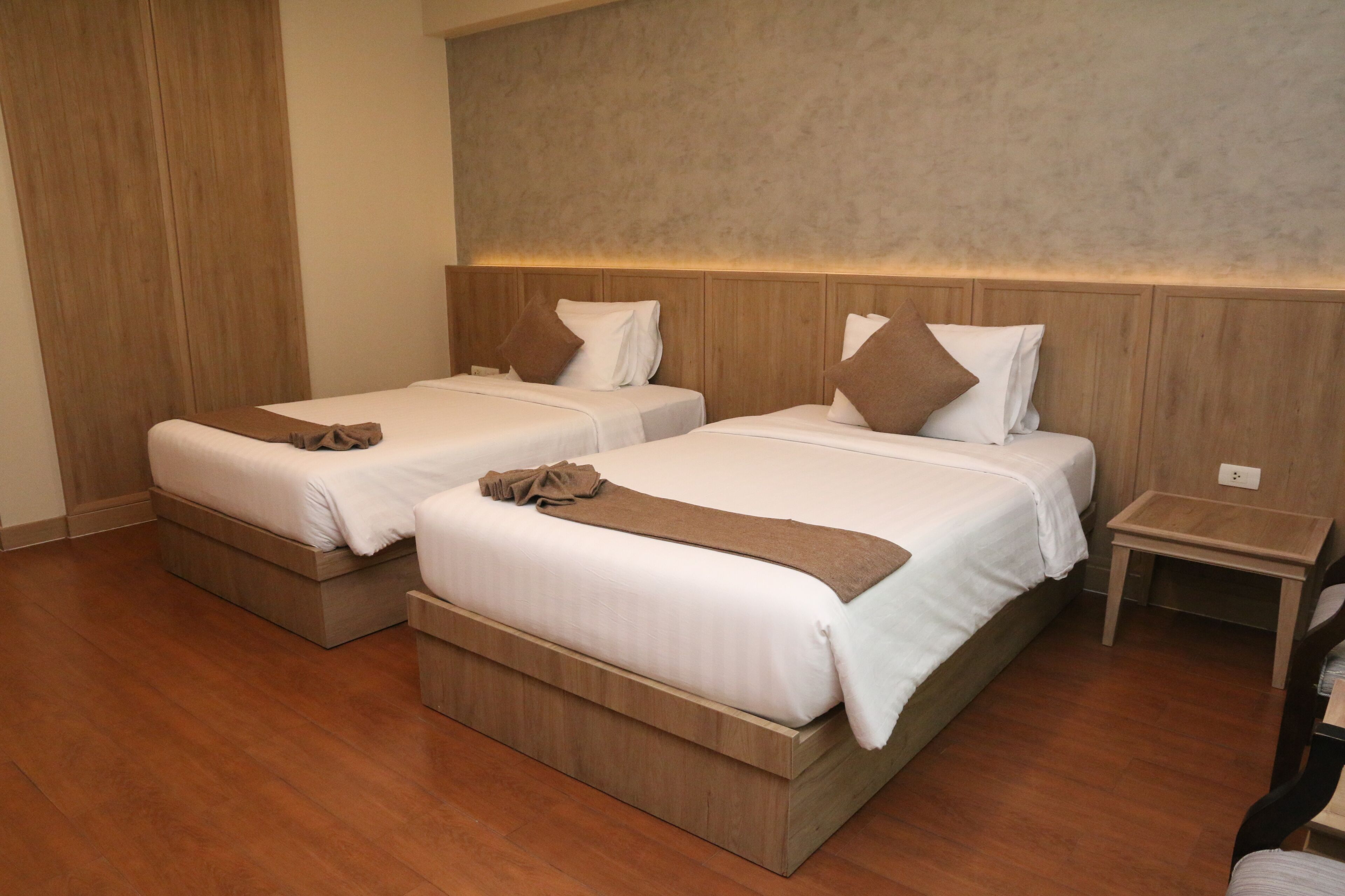 A hotel room with two beds, wooden headboards, a small bedside table, and a wooden floor.