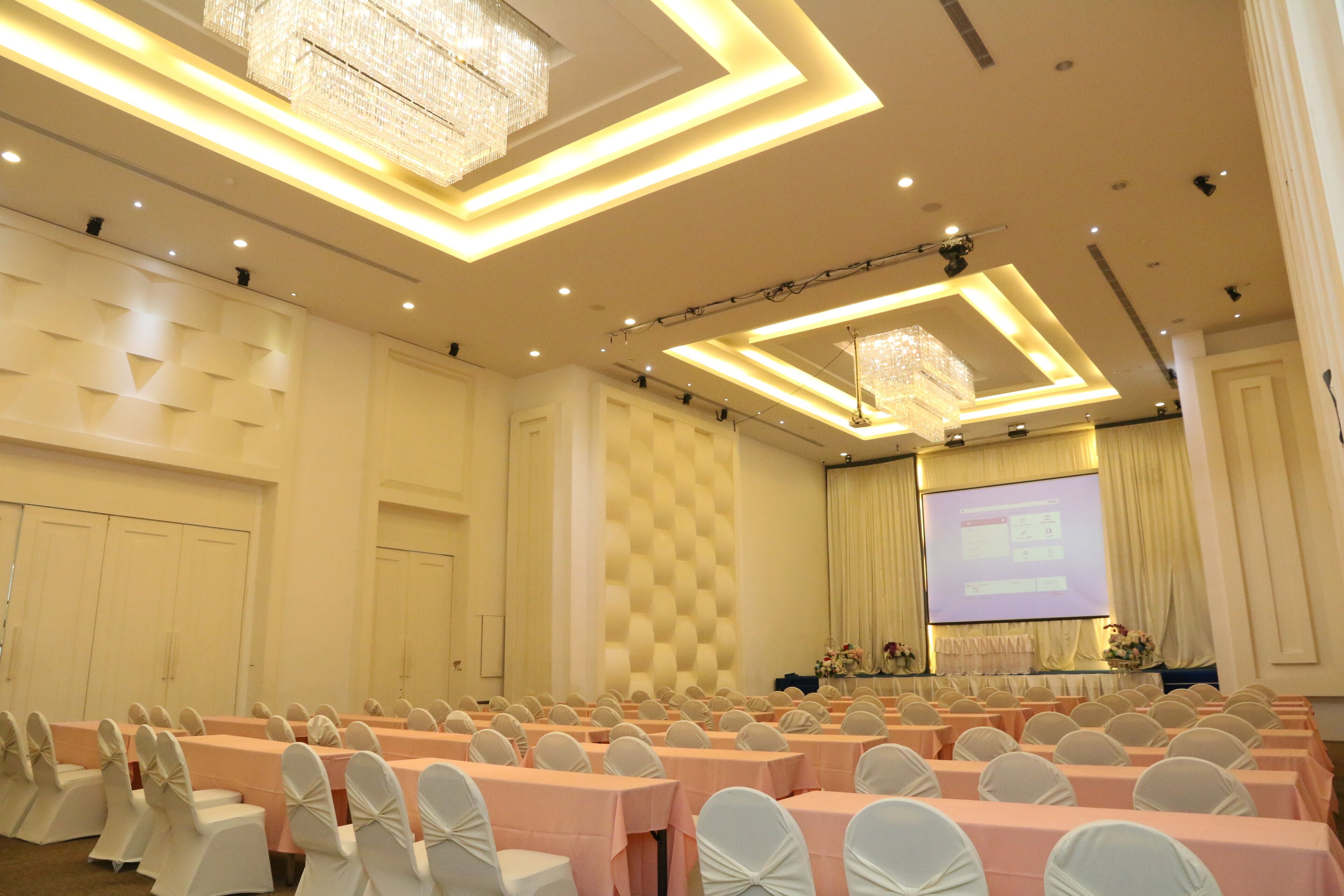 A conference room with rows of tables and chairs, a projector screen, and a stage with a floral arrangement.