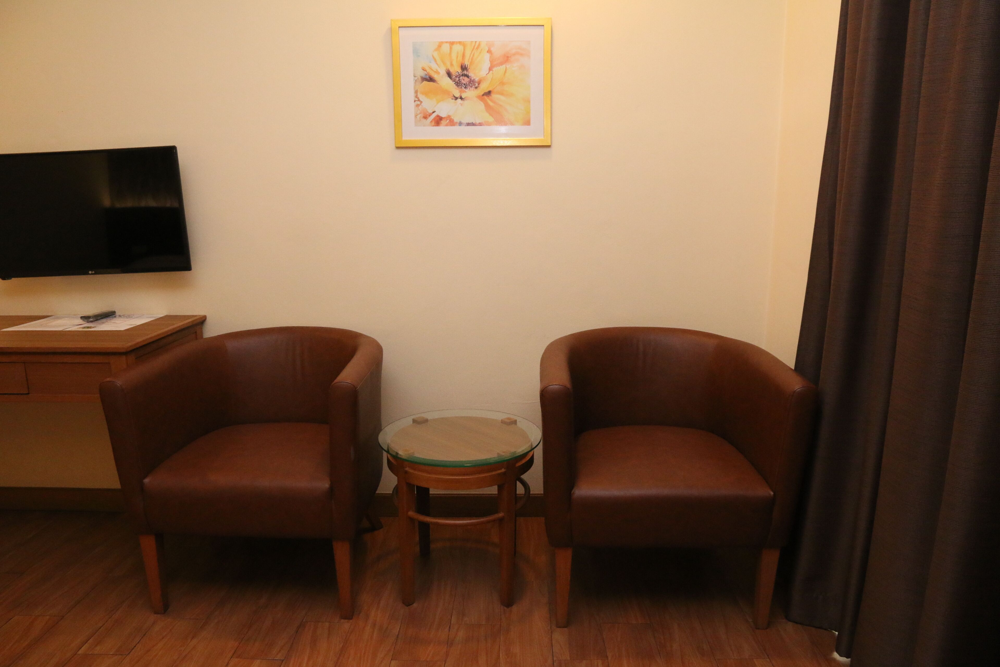 A room with two brown leather chairs, a small glass table, a flat-screen TV on a wooden stand, and a framed painting on the wall.