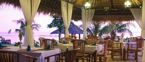 An outdoor dining area with tables and chairs, a thatched roof, and a view of the ocean.