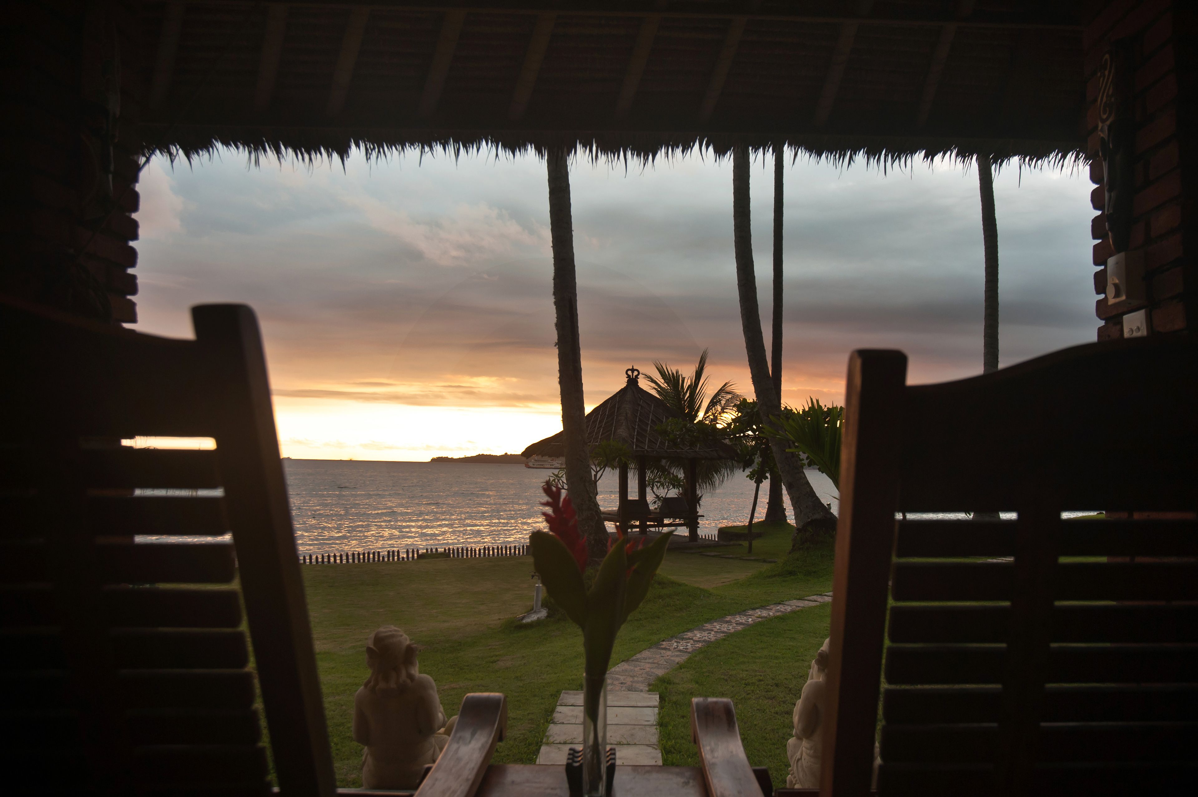 Une vue prise depuis un espace abrité, avec des chaises en bois, une structure à toit de chaume et des palmiers surplombant une étendue d’eau au coucher du soleil.