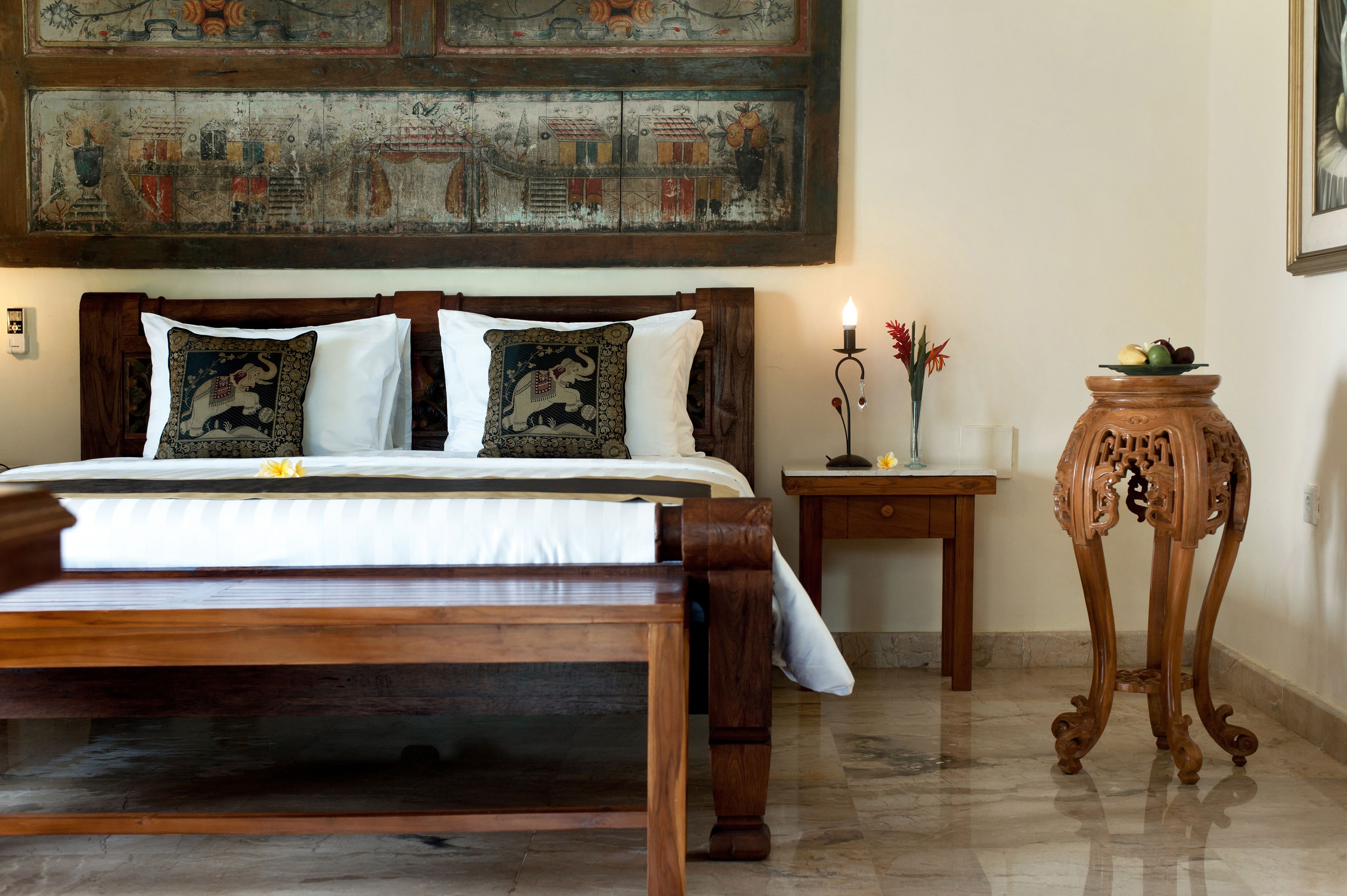 Un dormitorio con una cama de madera, dos almohadas decorativas, una mesita de noche con una vela y una pequeña mesa con fruta.